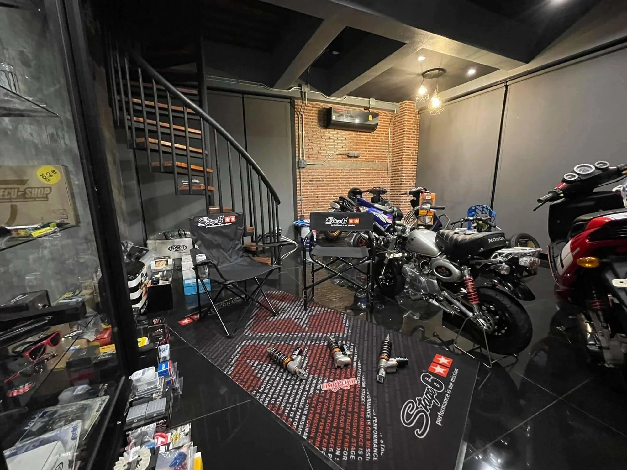 Motorsports gear shop with motorcycles, racing parts, and accessories displayed on shelves and floor, brick wall backdrop, black and gray interior, and overhead lighting.