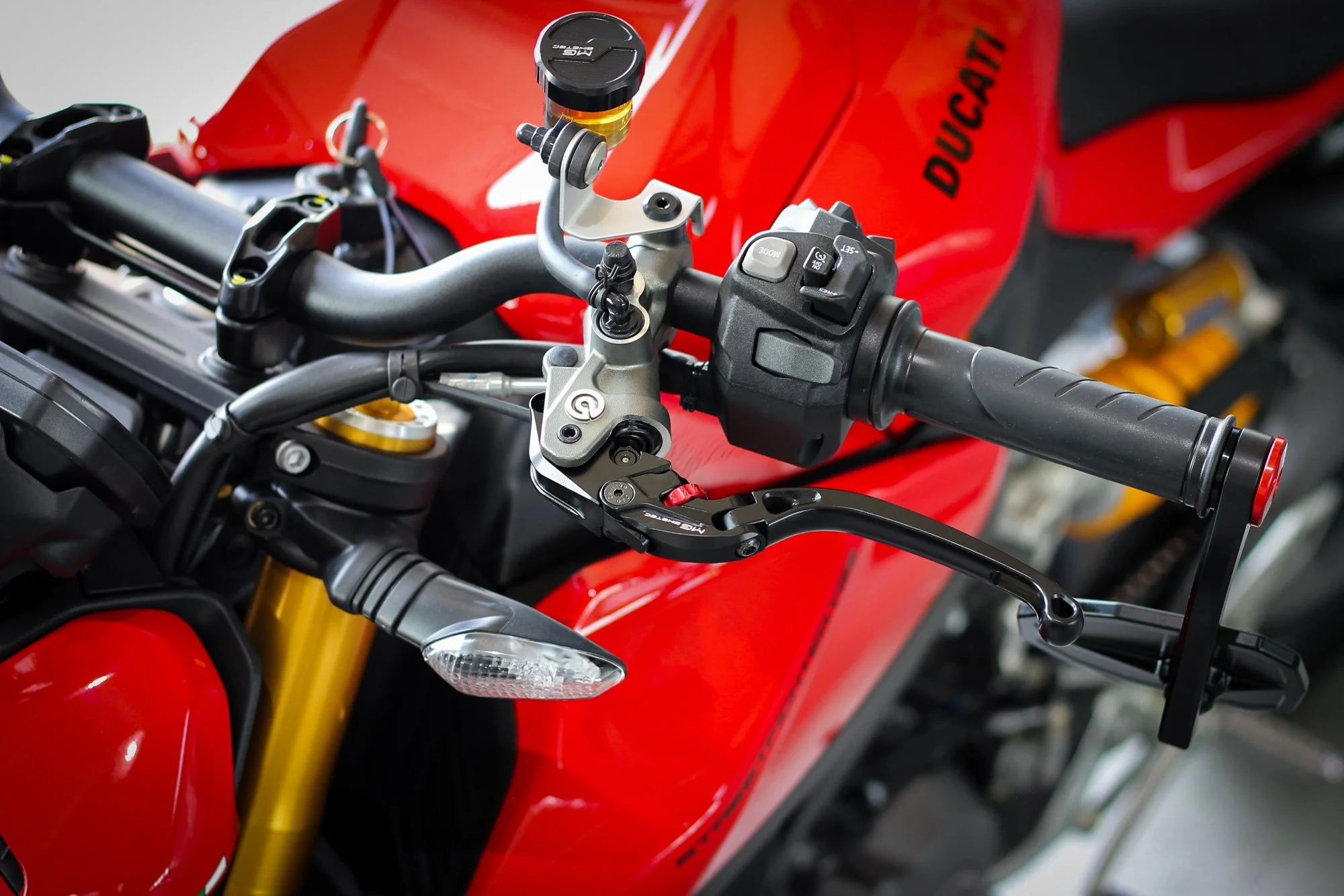 Close-up of a red Ducati motorcycle handlebar, showing the throttle grip, control switches, and a small tank with a black cap, with parts of the front suspension and headlight visible.