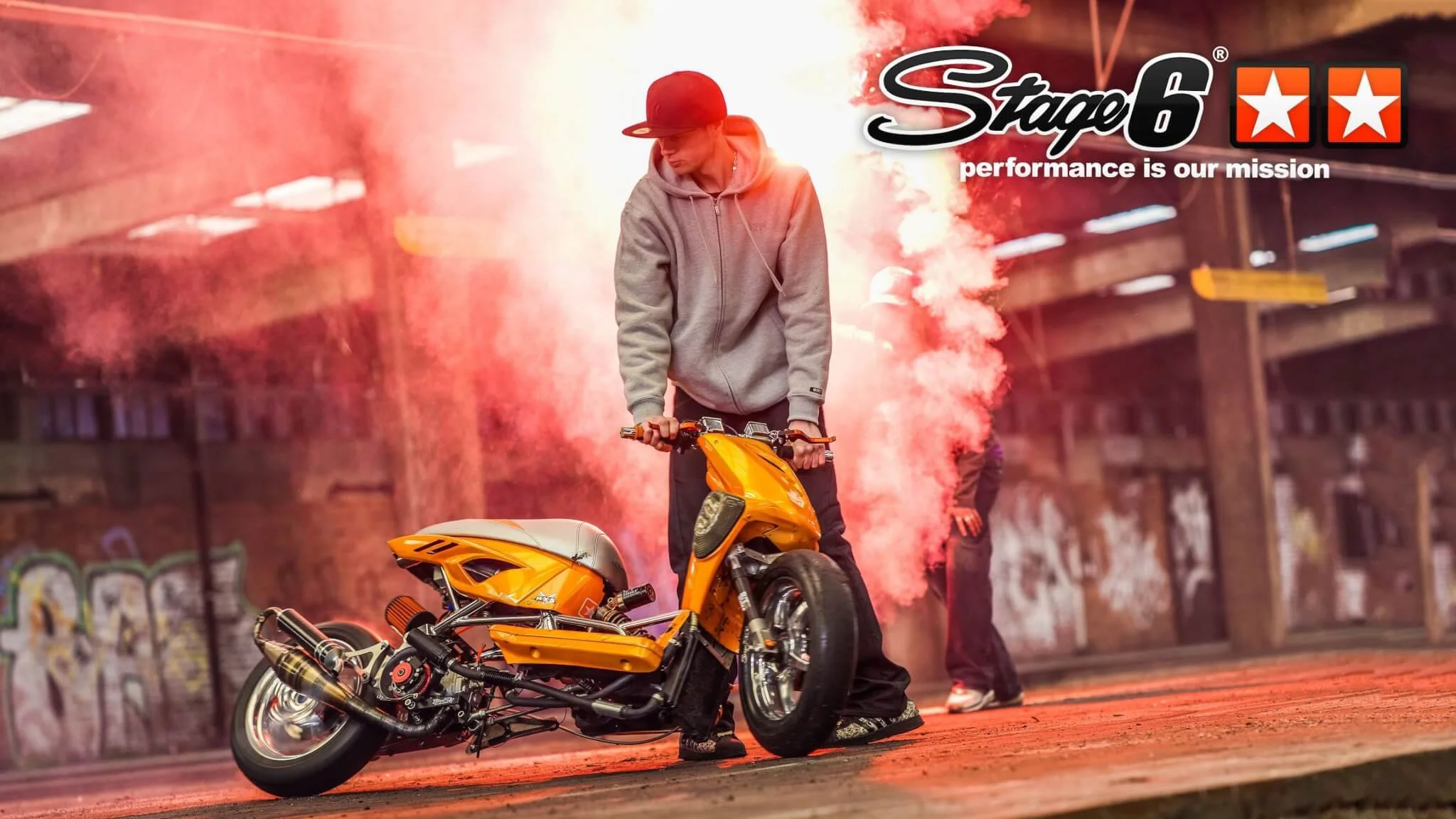 A young man in a gray hoodie and cap sitting on a bright yellow motorcycle in an urban setting with graffiti on the walls, with red smoke and sparks around, and another person in the background.
