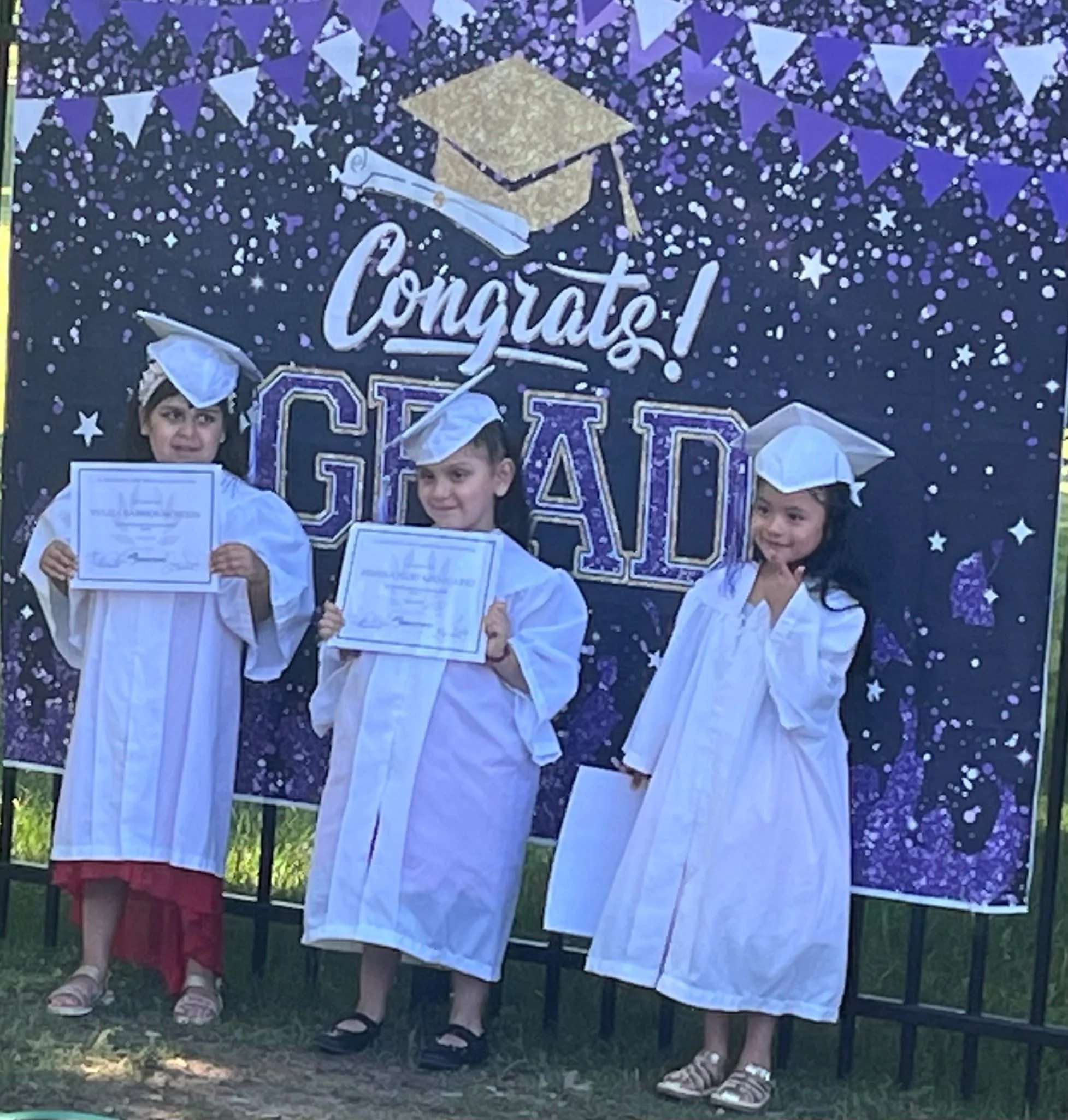 Three young girls dressed in graduation caps and gowns holding diplomas, standing in front of a backdrop that says 'Congrats! Grad' with a graduation cap graphic and star decorations.