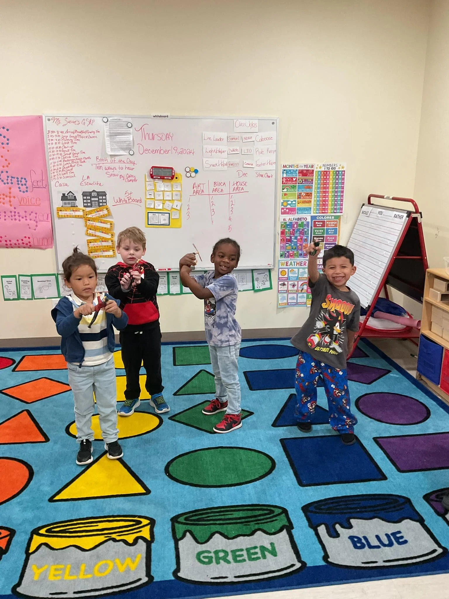 Four young children standing on a colorful classroom rug, raising their hands and smiling, in front of a whiteboard with educational posters and charts.