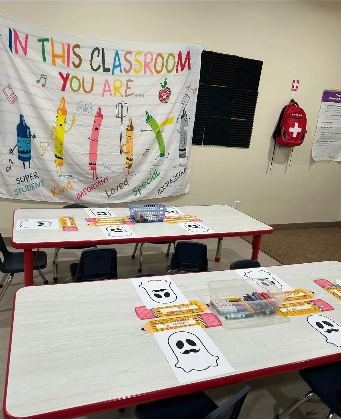 Classroom with tables decorated for Halloween, featuring paper ghost shapes, crayons, and name tags. A colorful banner on the wall reads 'In this classroom, you are...' with words describing students.