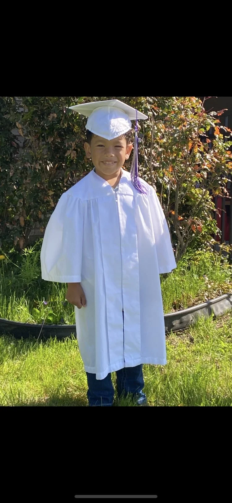 Young boy in a white graduation cap and gown, smiling outdoors on grass with bushes in the background.