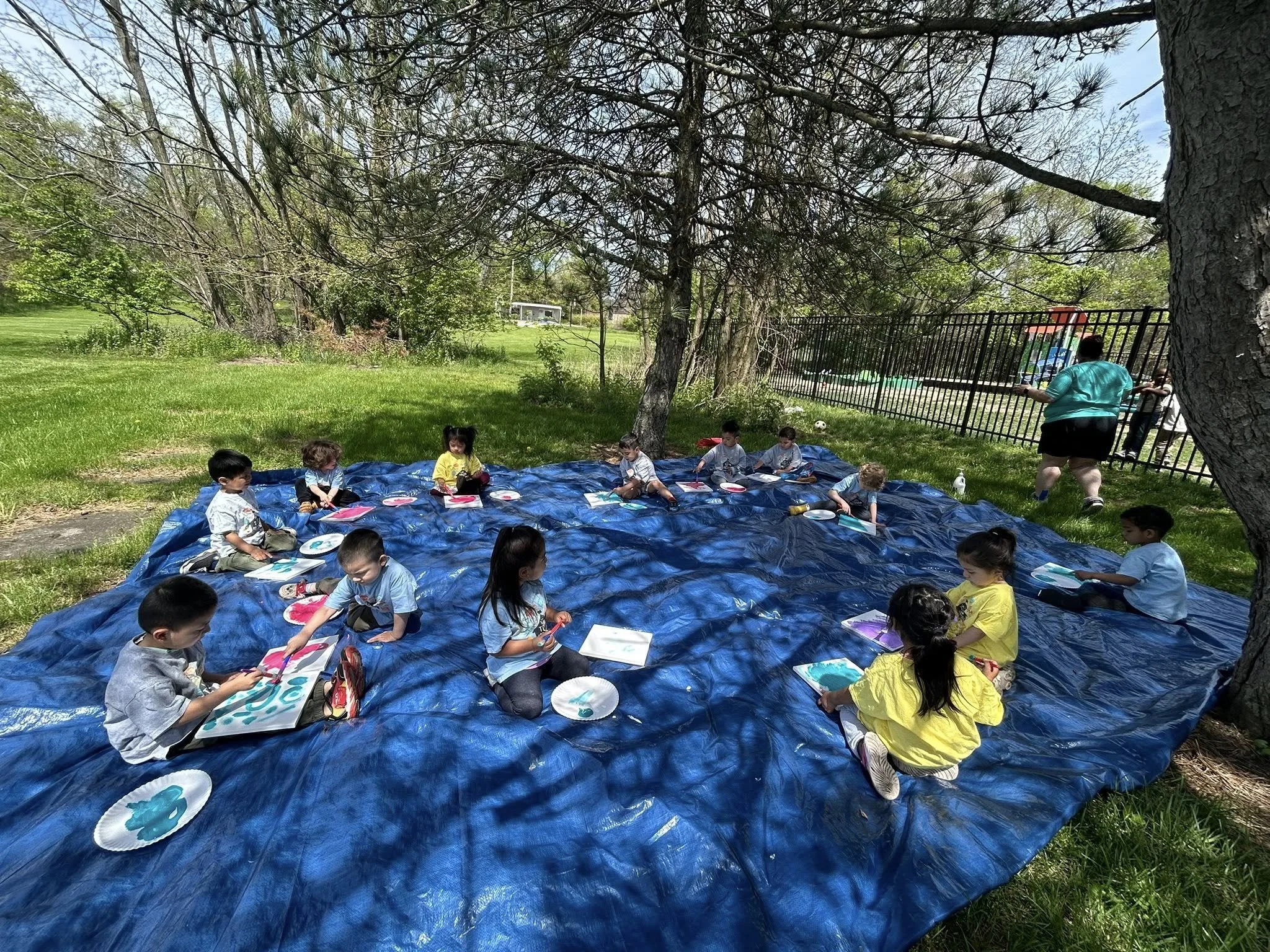 Children sitting on a large blue tarp under trees, painting on paper with plates of paint, outdoors near a fenced playground.