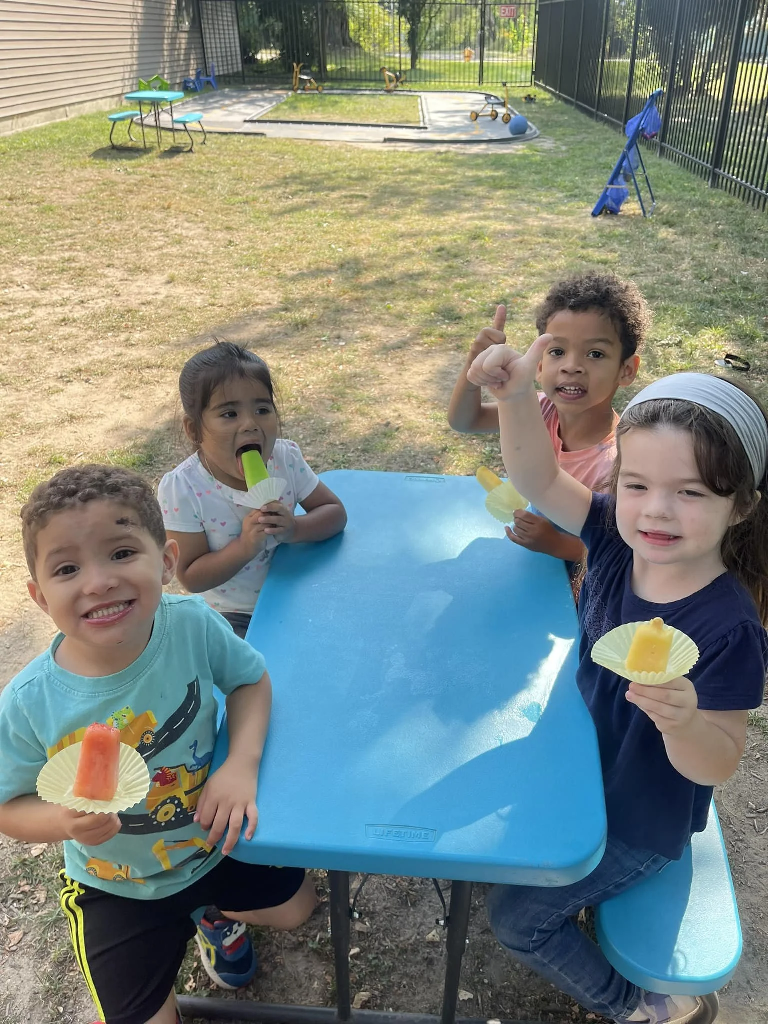 Four children at an outdoor table enjoying popsicles the kids are smiling and excited, with some raising their hands, and a playground with a sandbox and toys is visible in the background.