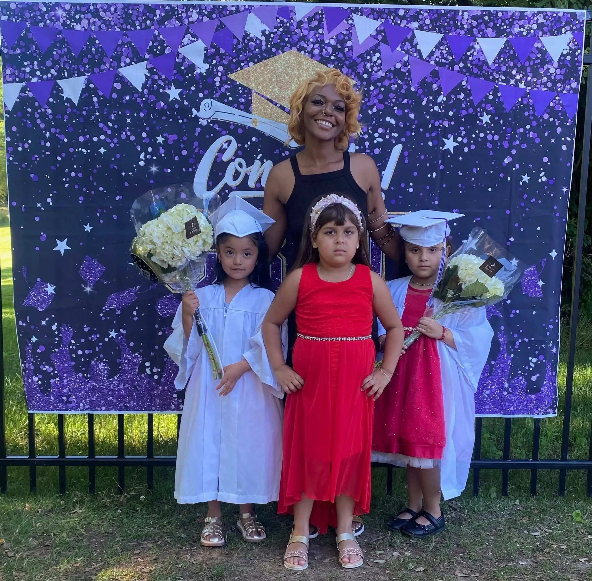 A woman with blonde curly hair in a black dress stands behind three young girls dressed for a graduation, two in white caps and gowns holding bouquets, and one in a red dress. They are standing in front of a purple graduation-themed backdrop with con