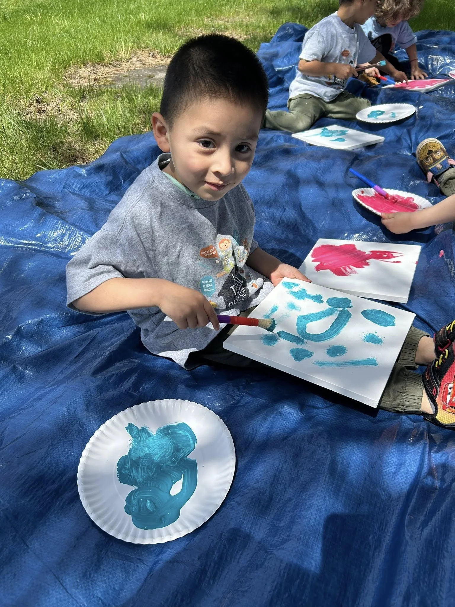 A young boy sitting on a blue tarp outdoors, painting on a paper with blue paint. There are other children in the background also painting. The boy is holding a paintbrush and looking at the camera.
