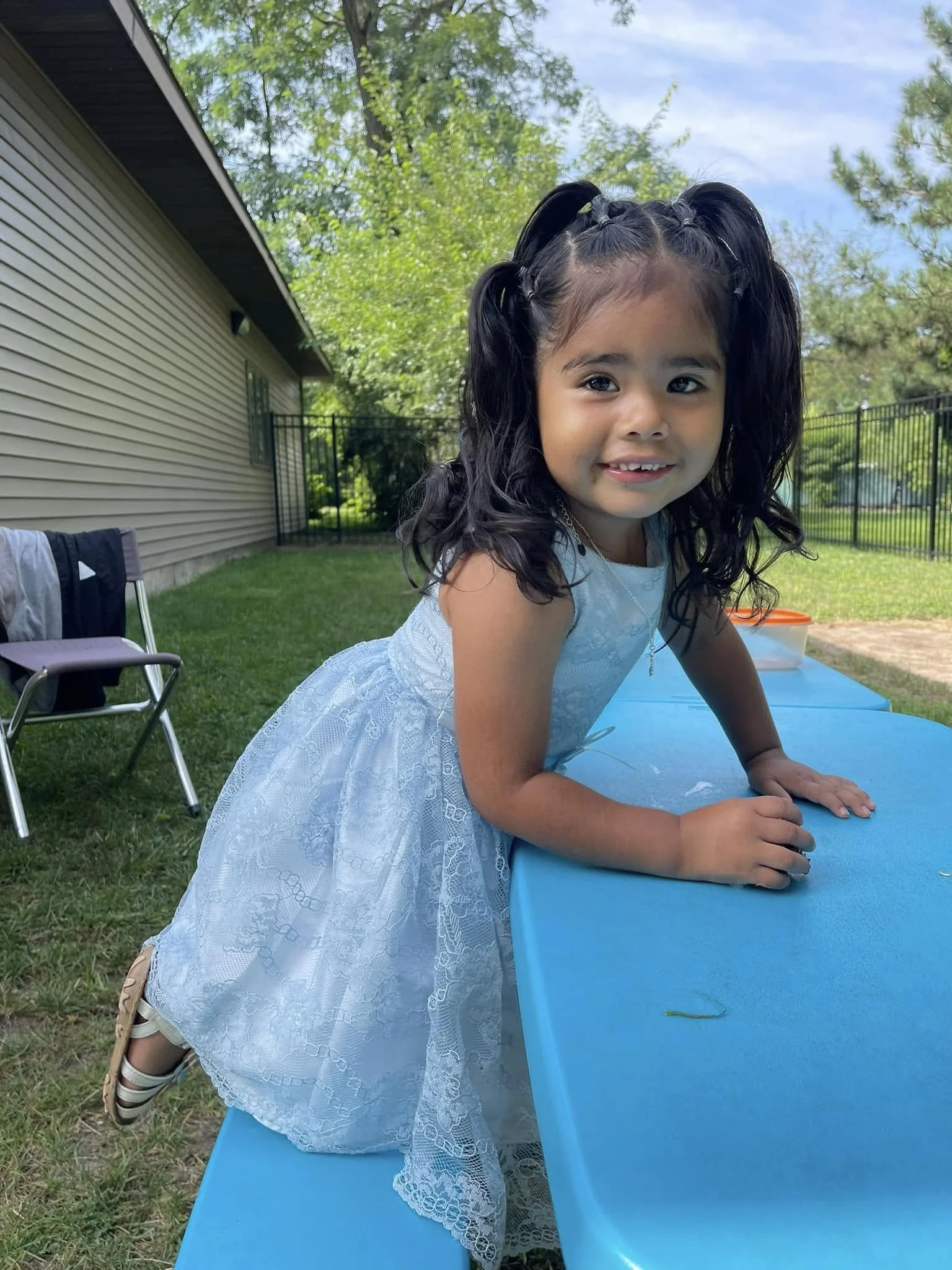 A young girl with dark, curly hair in pigtails, wearing a white lace dress, leaning on a blue table outdoors, smiling at the camera. There are trees, a house, and a black fence in the background.