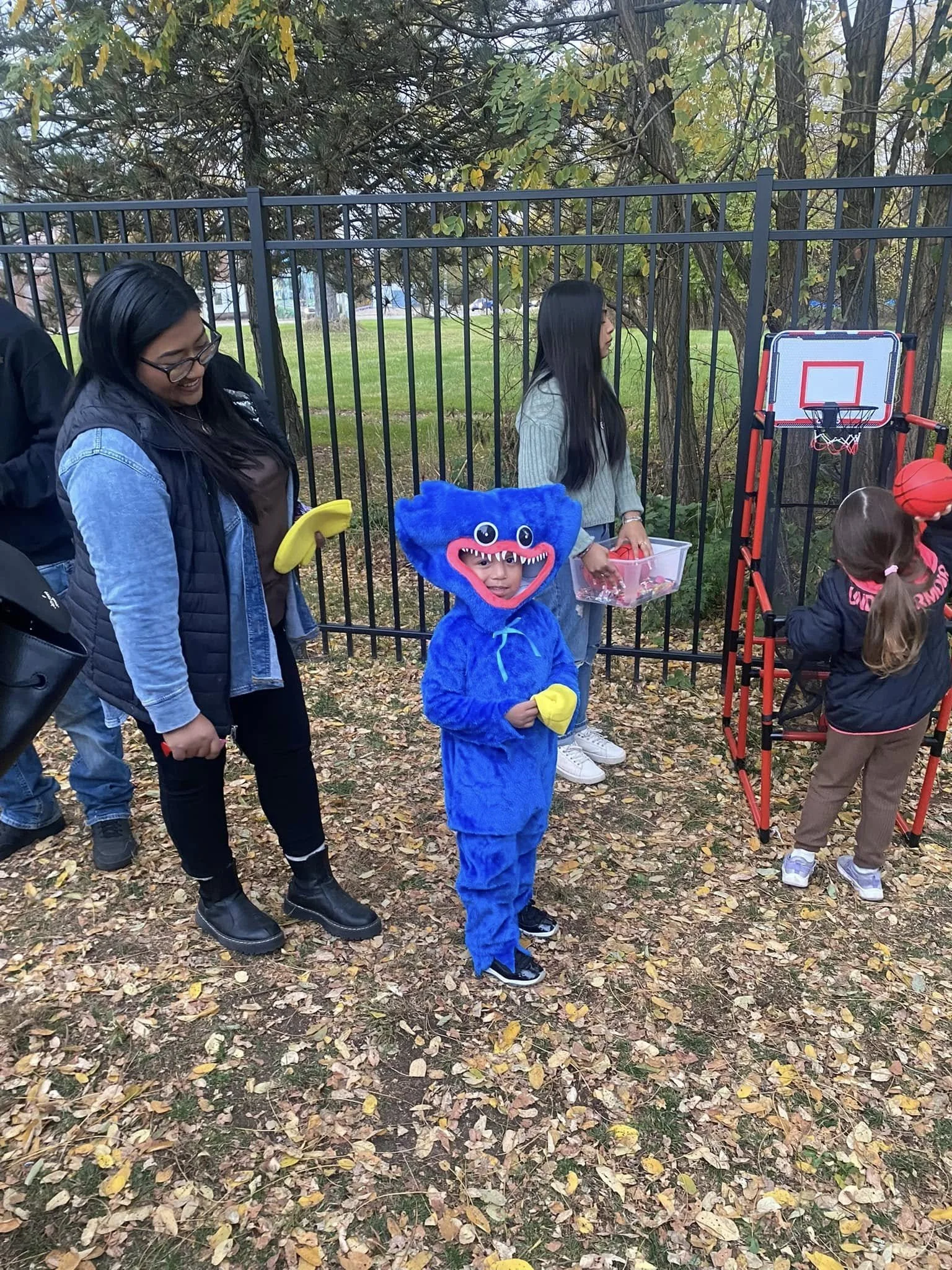 Children and adults outside playing with a basketball and a mini basketball hoop, dressed in fall clothing, surrounded by fallen leaves, with trees and a metal fence in the background.