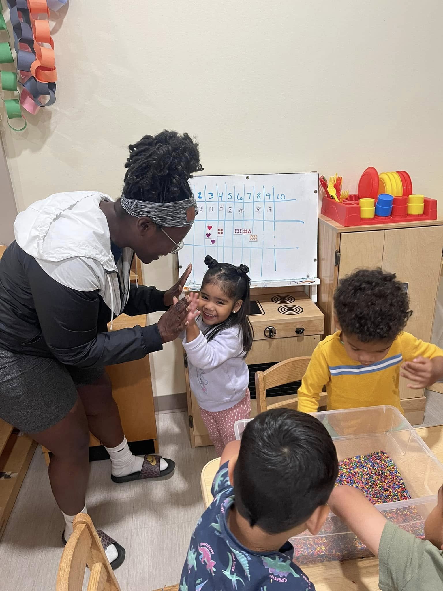 A woman giving a high five to a young girl in a classroom with three children, a whiteboard with numbers and animals, and colorful toys.