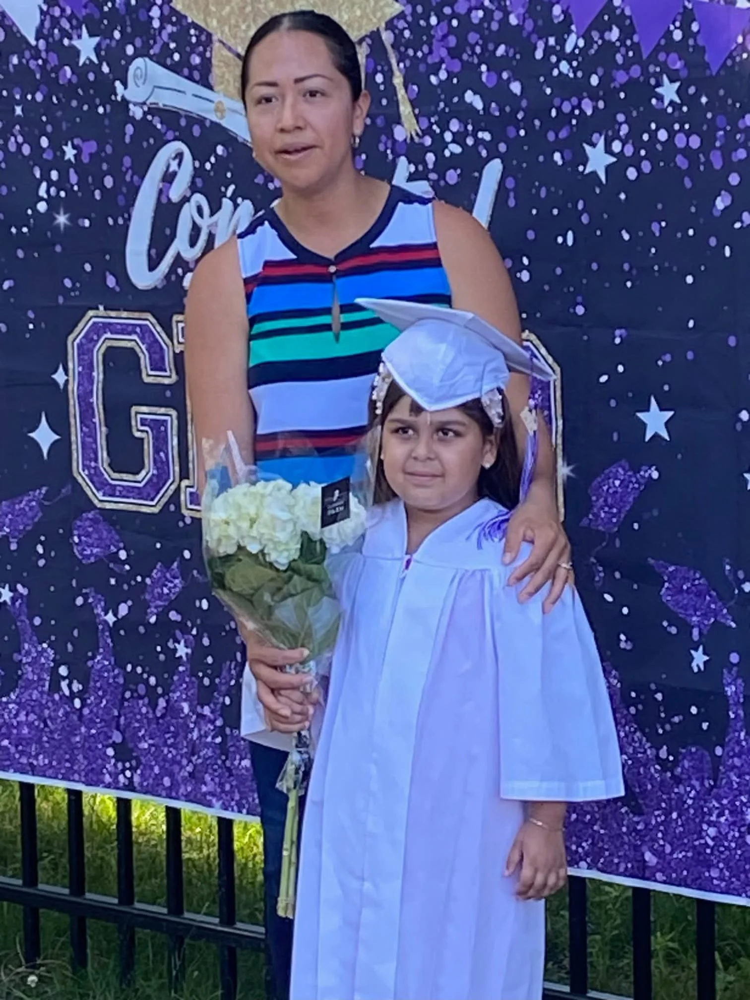 A young girl dressed in a white graduation gown and cap holding a bouquet of white flowers standing next to an adult woman in a striped sleeveless top, with a purple celebratory backdrop featuring stars and the word 'Congratulations'.