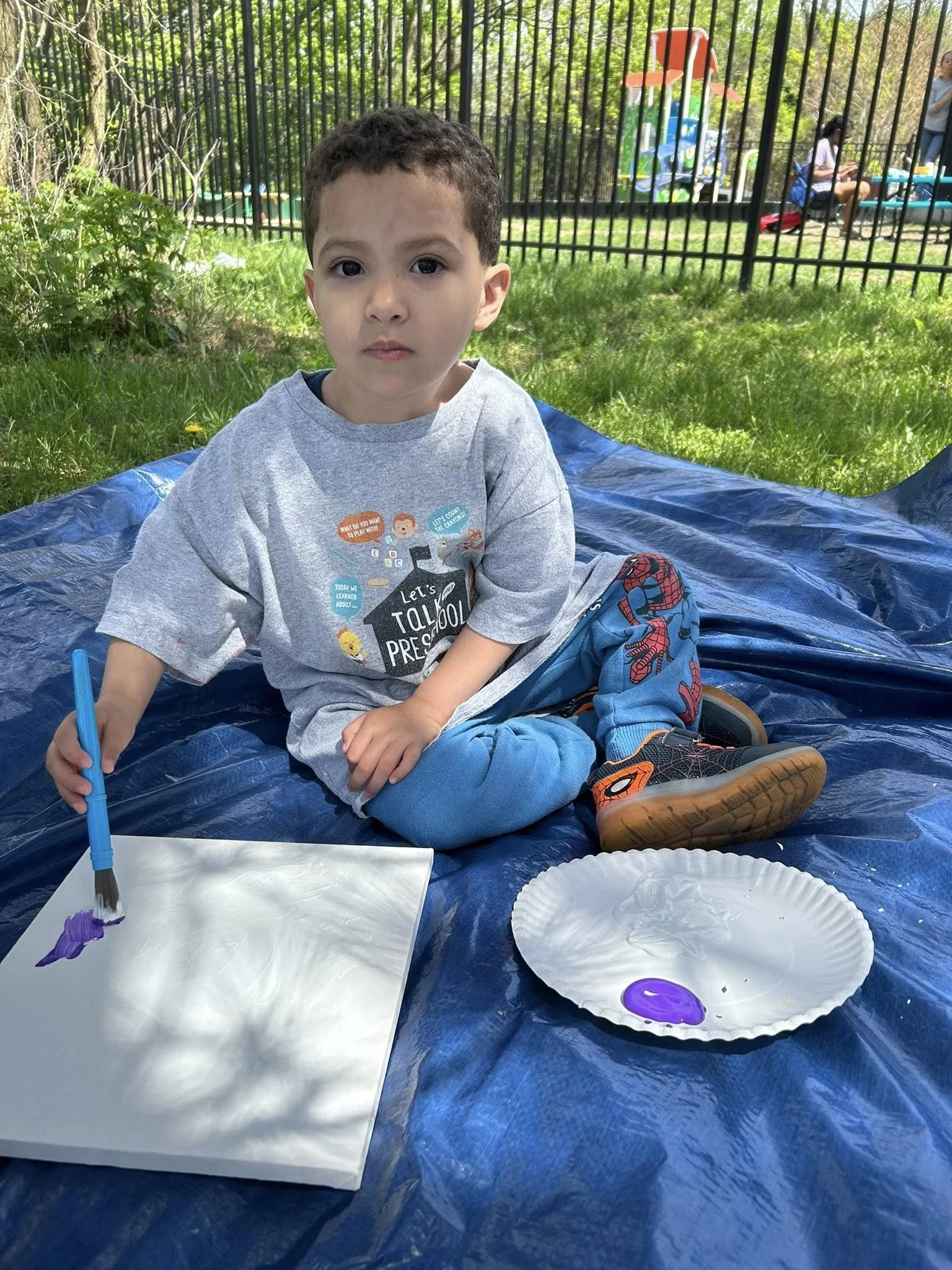 A young boy sitting on a blue tarp outdoors, painting on a piece of paper with purple paint. There is a paper plate with purple paint beside him. In the background, there are trees, a metal fence, and people near a playground.