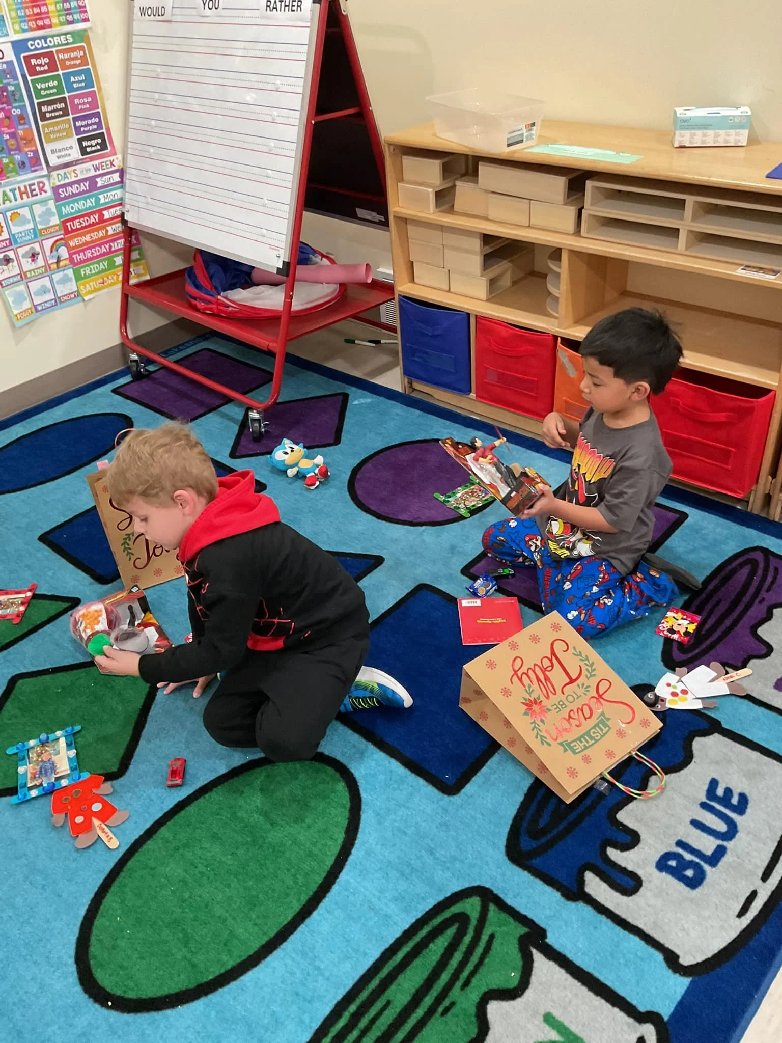 Two boys sitting on a colorful classroom rug filled with toys and holiday gift bags, opening presents, with educational posters and storage units in the background.