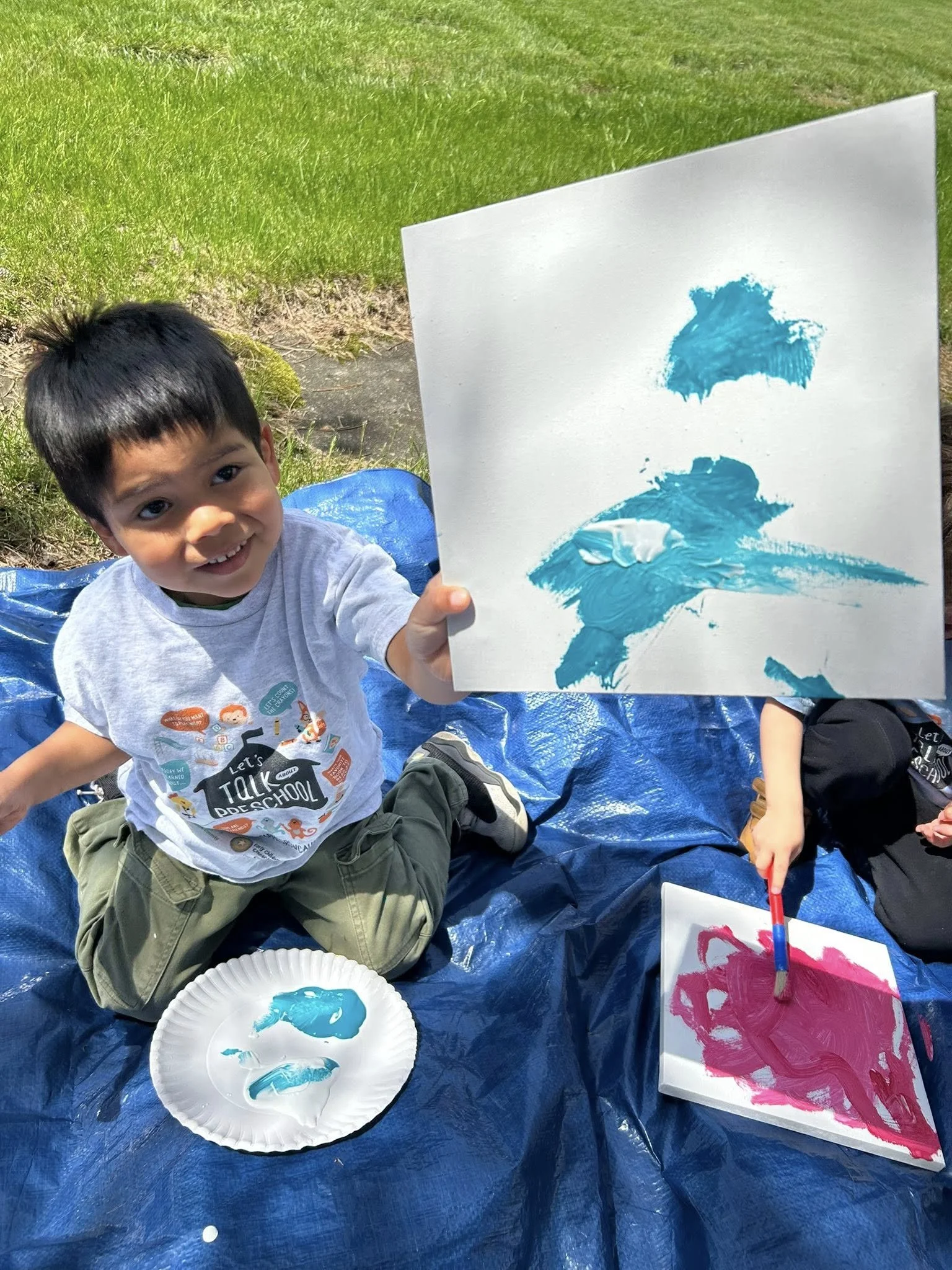 A young boy sitting on a blue tarp outdoors, holding up a piece of paper with blue paint on it. He is smiling and wearing a grey T-shirt with colorful designs. There is a paper plate with blue paint and a brush nearby. Another person is partially vis