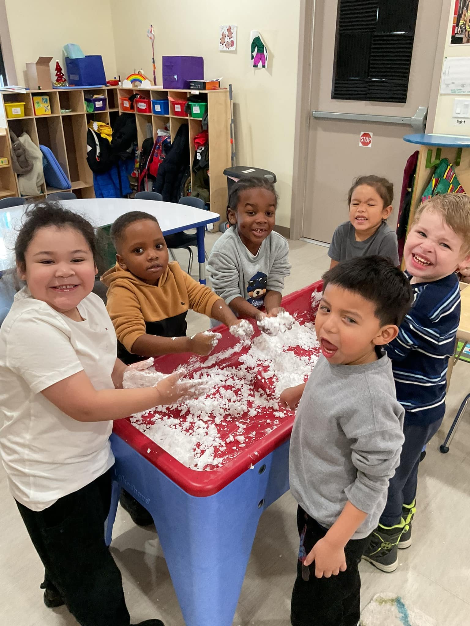 Six children playing with and touching a large bin of snow or white powder in a classroom.