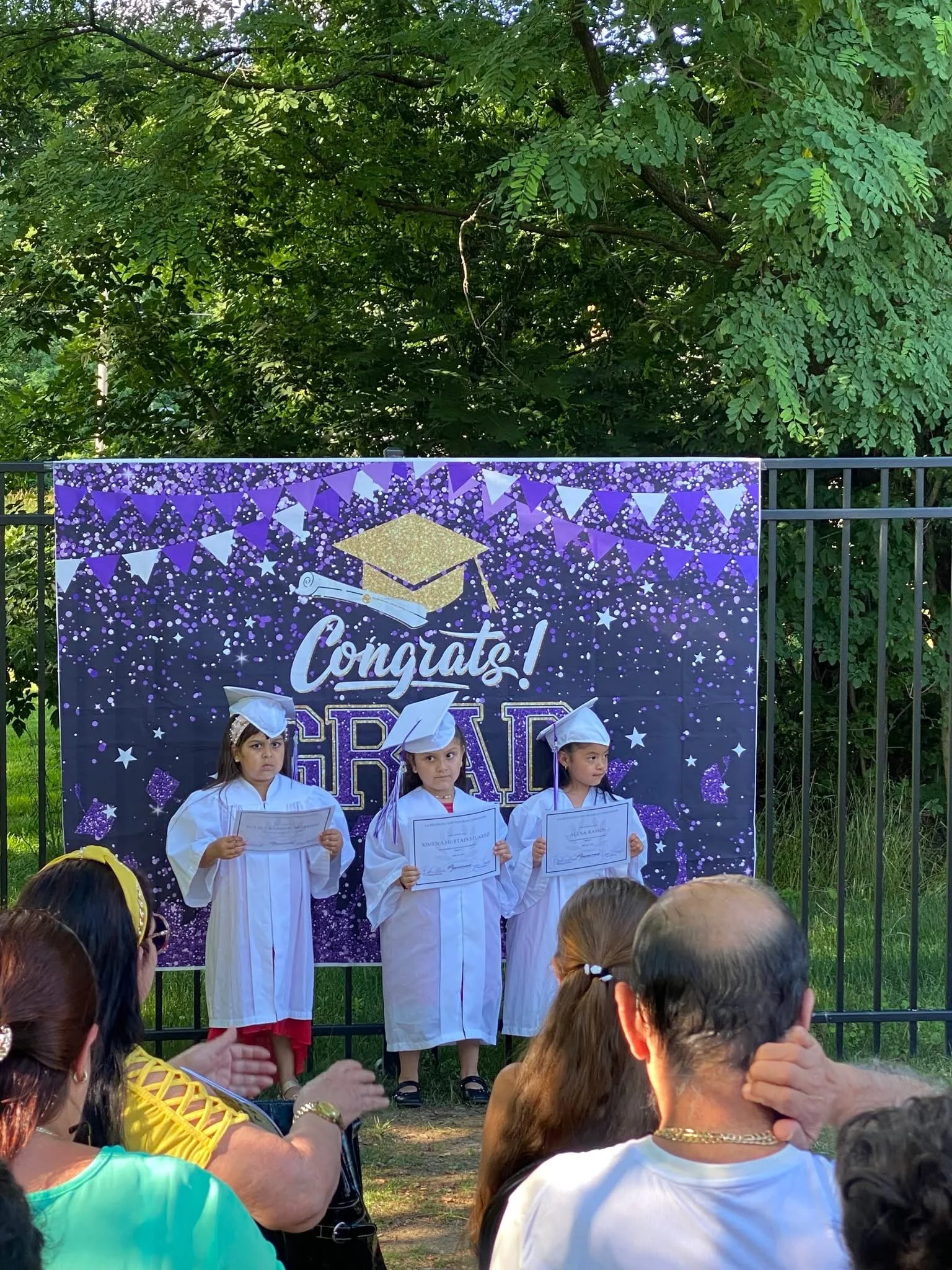 Three young children wearing white graduation gowns and caps stand on a stage holding diplomas during a graduation ceremony outdoors. Behind them is a purple decorative backdrop with a graduation cap graphic, the words "Congrats! GRAD," and celebrato