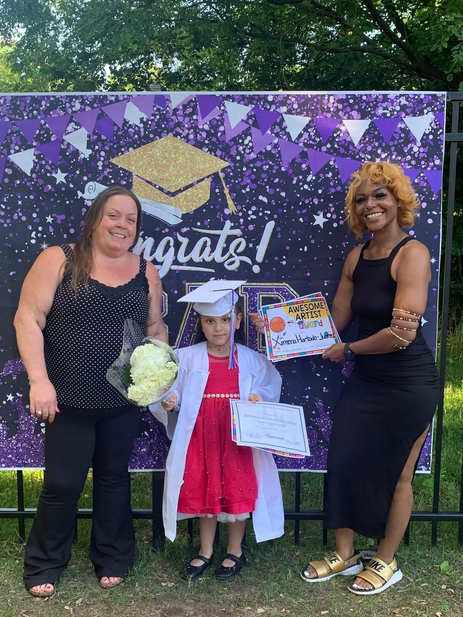 A young girl wearing a white graduation gown and cap holds a diploma, standing between two women who are celebrating her graduation. The woman on the left has long hair and is wearing a black polka dot blouse, while the woman on the right has curly b
