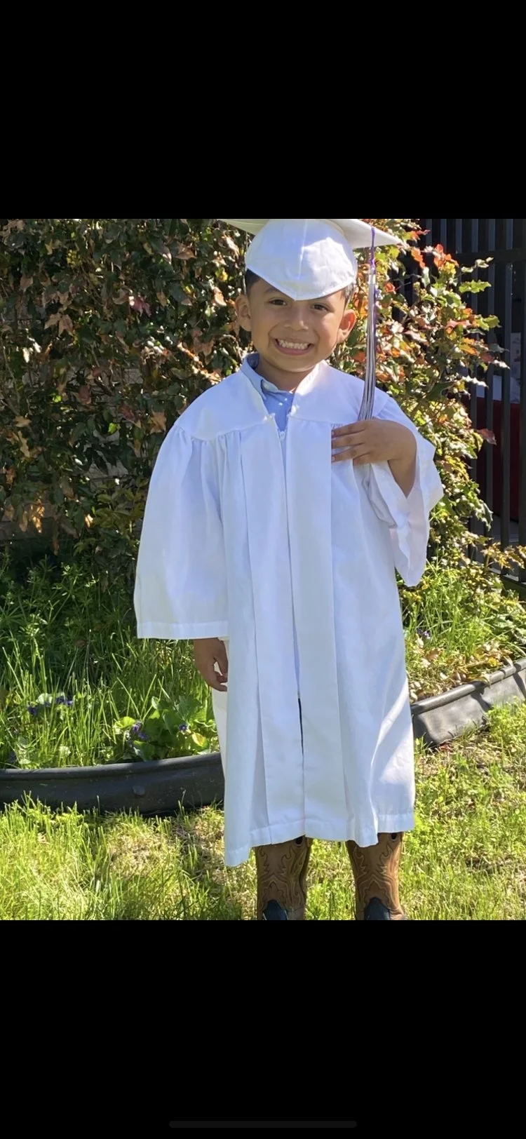 A young boy dressed in a white graduation gown and cap, holding a small umbrella, standing outdoors in front of green bushes and grass, smiling.