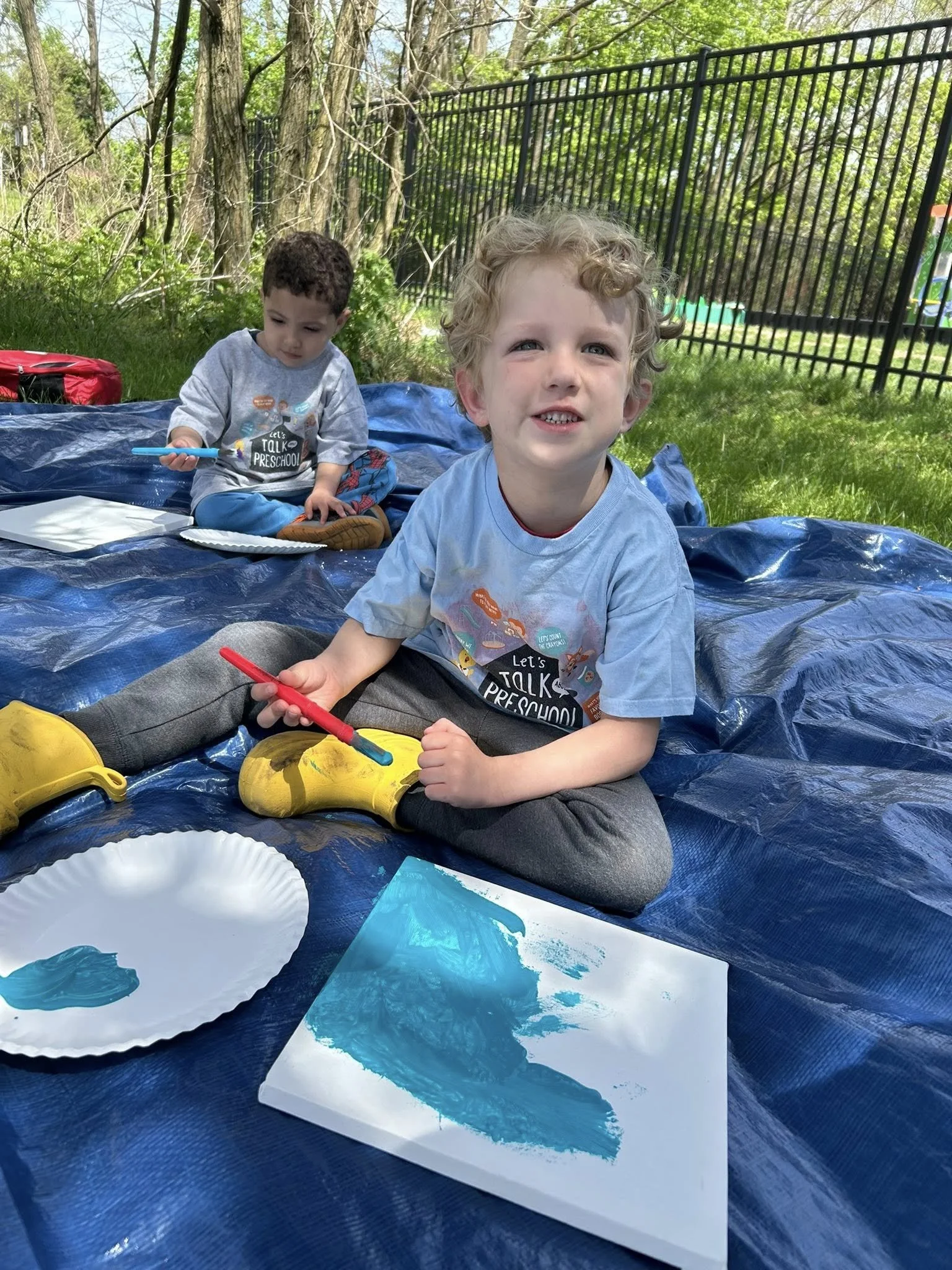 Two young children painting outdoors on a blue tarp, with one child holding a paintbrush and paper, and a nearby plate with blue paint; trees and a black fence in the background.