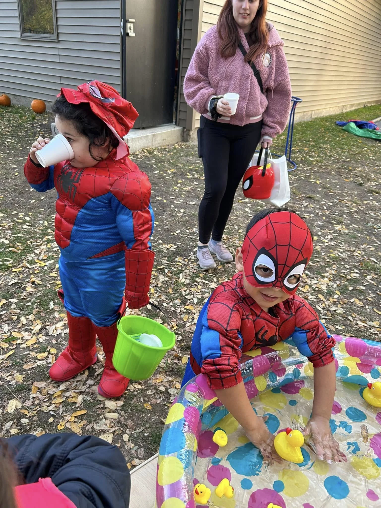 Children dressed as Spider-Man at a fall outdoor party, with one child playing in a small inflatable pool with rubber duckies and another child drinking from a cup, while an adult woman stands nearby holding a white cup and a Disney-themed bag.