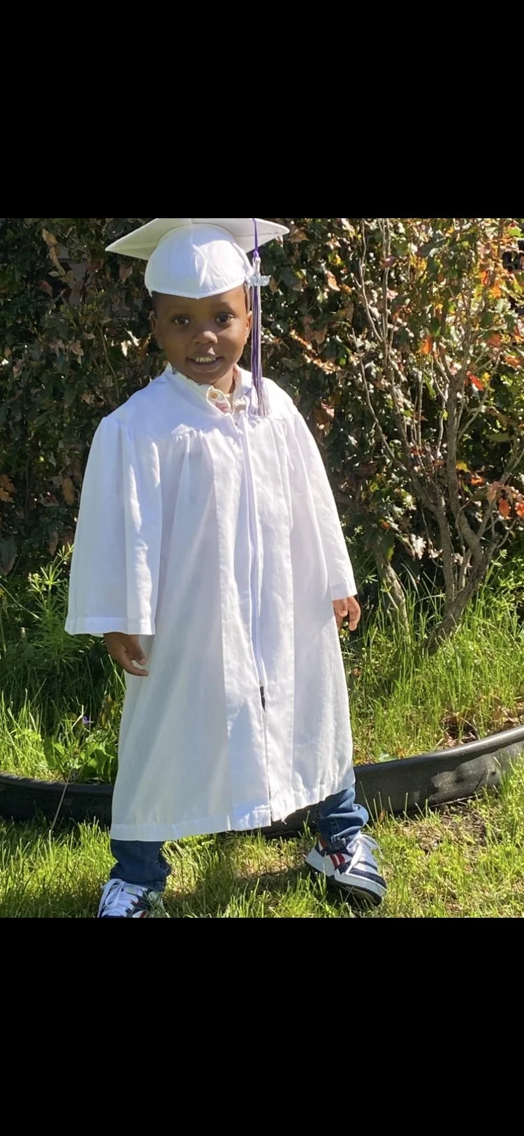 A young boy dressed in a white graduation gown and cap, standing on grass in front of bushes, wearing sneakers.