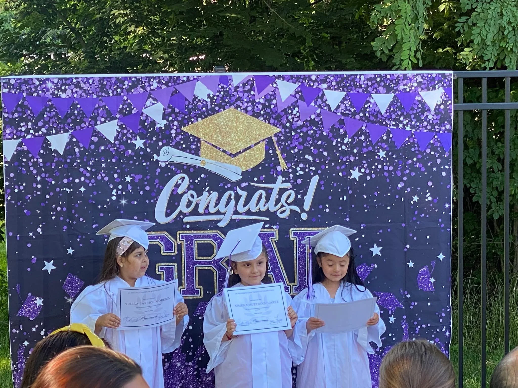 Three young girls dressed in white graduation caps and gowns holding certificates, standing in front of a decorated backdrop with the words 'Congrats! GRAD' and a graduation cap graphic, at an outdoor graduation ceremony.