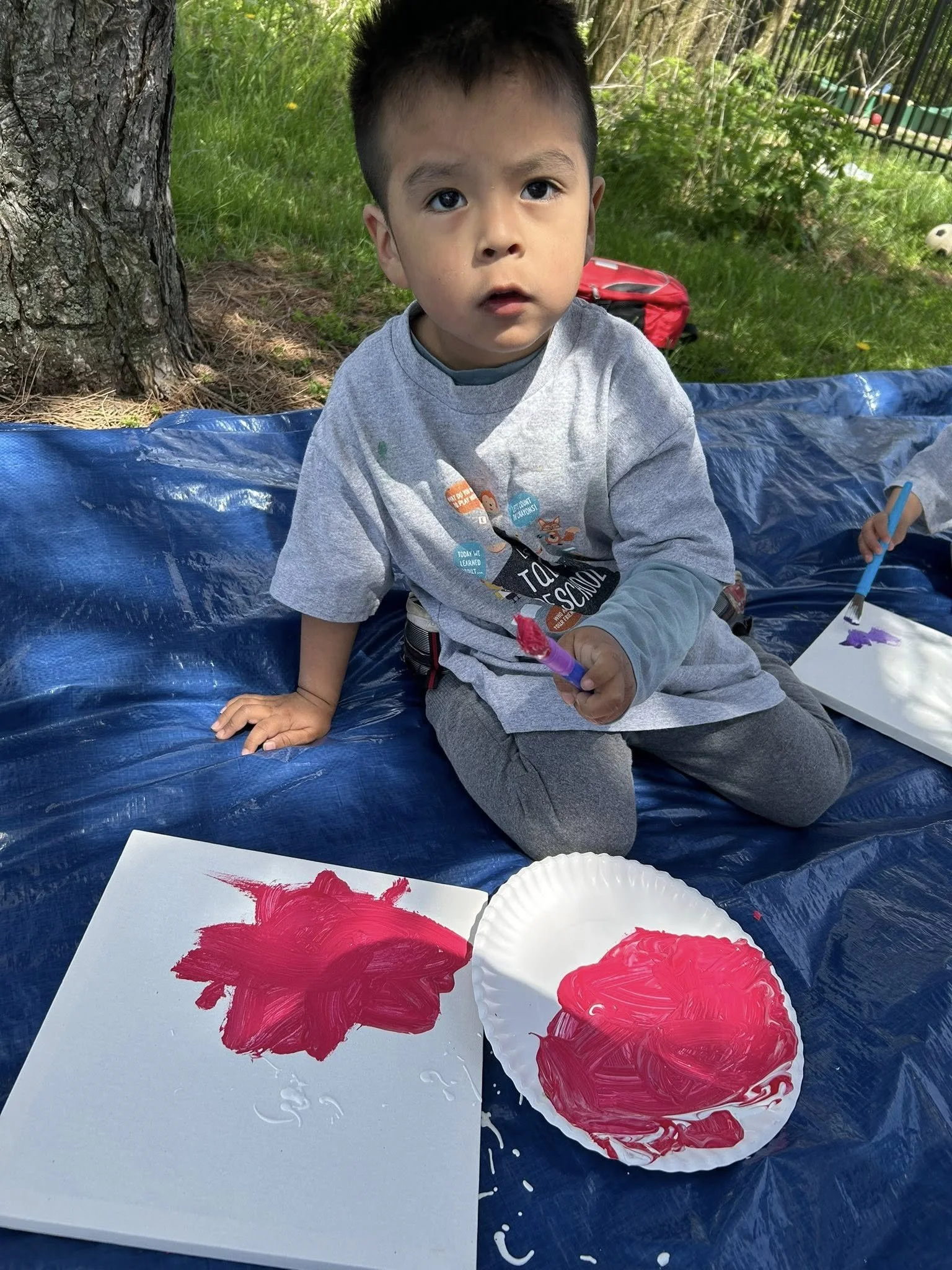A young boy sitting outdoors on a blue tarp, holding a paintbrush, with a paper plate and paper in front of him painted with red and purple colors.