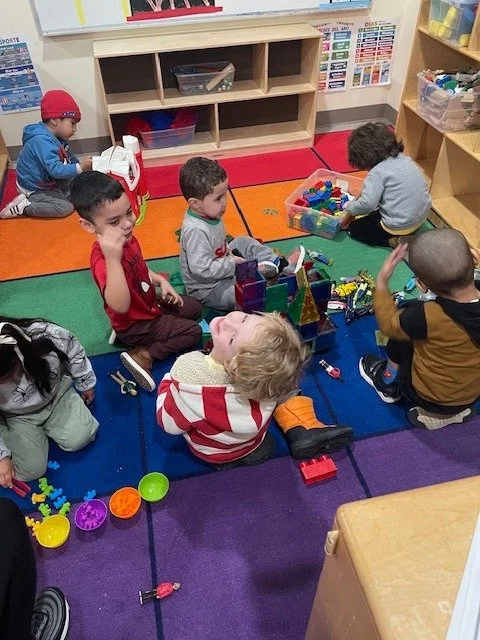 Children playing with toys in a colorful classroom, sitting on a rainbow-colored rug.