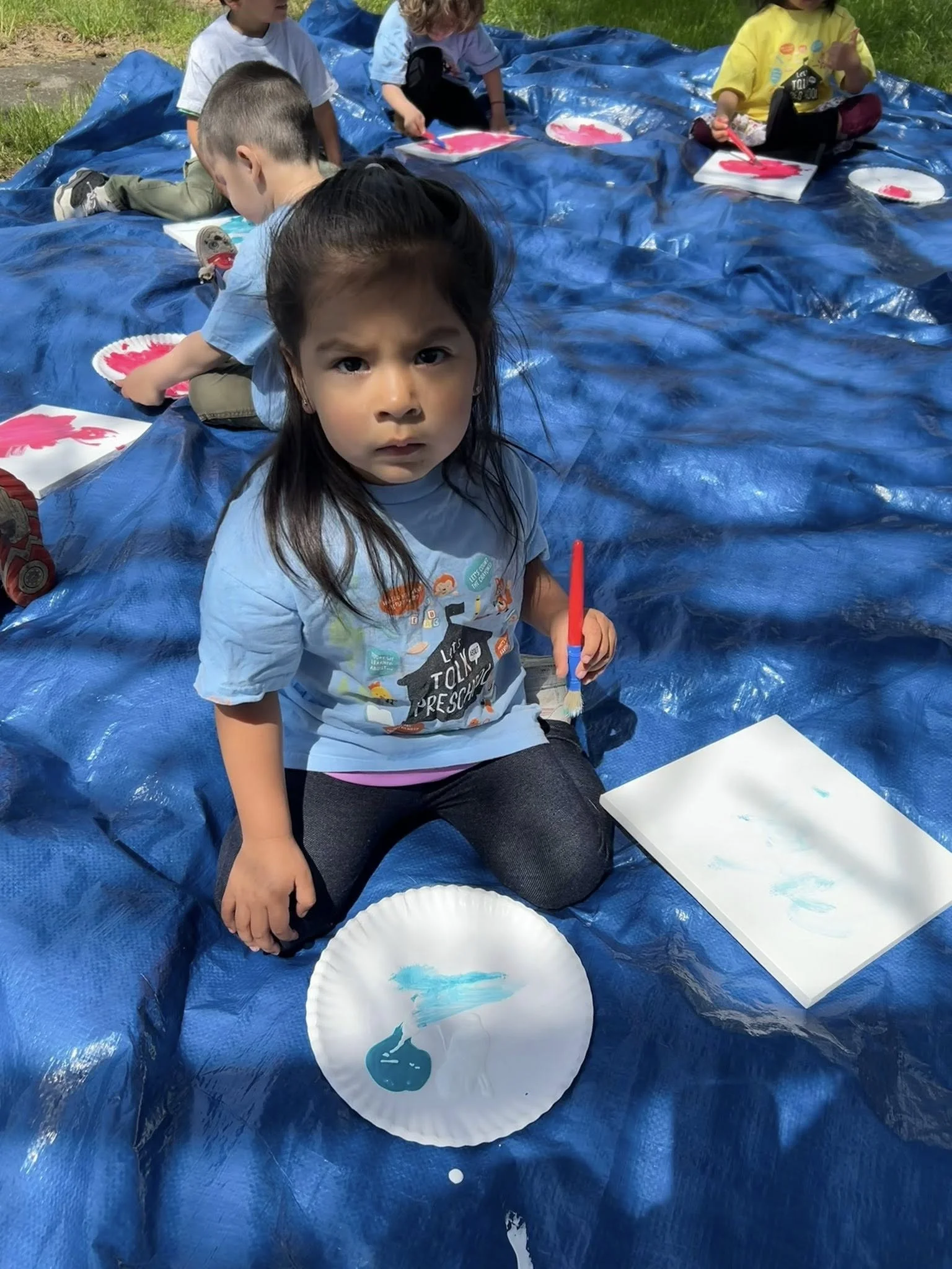 Girl sitting on a blue tarp outside, holding a red paintbrush with blue paint on a paper plate and a white sheet of paper, with children in the background painting.