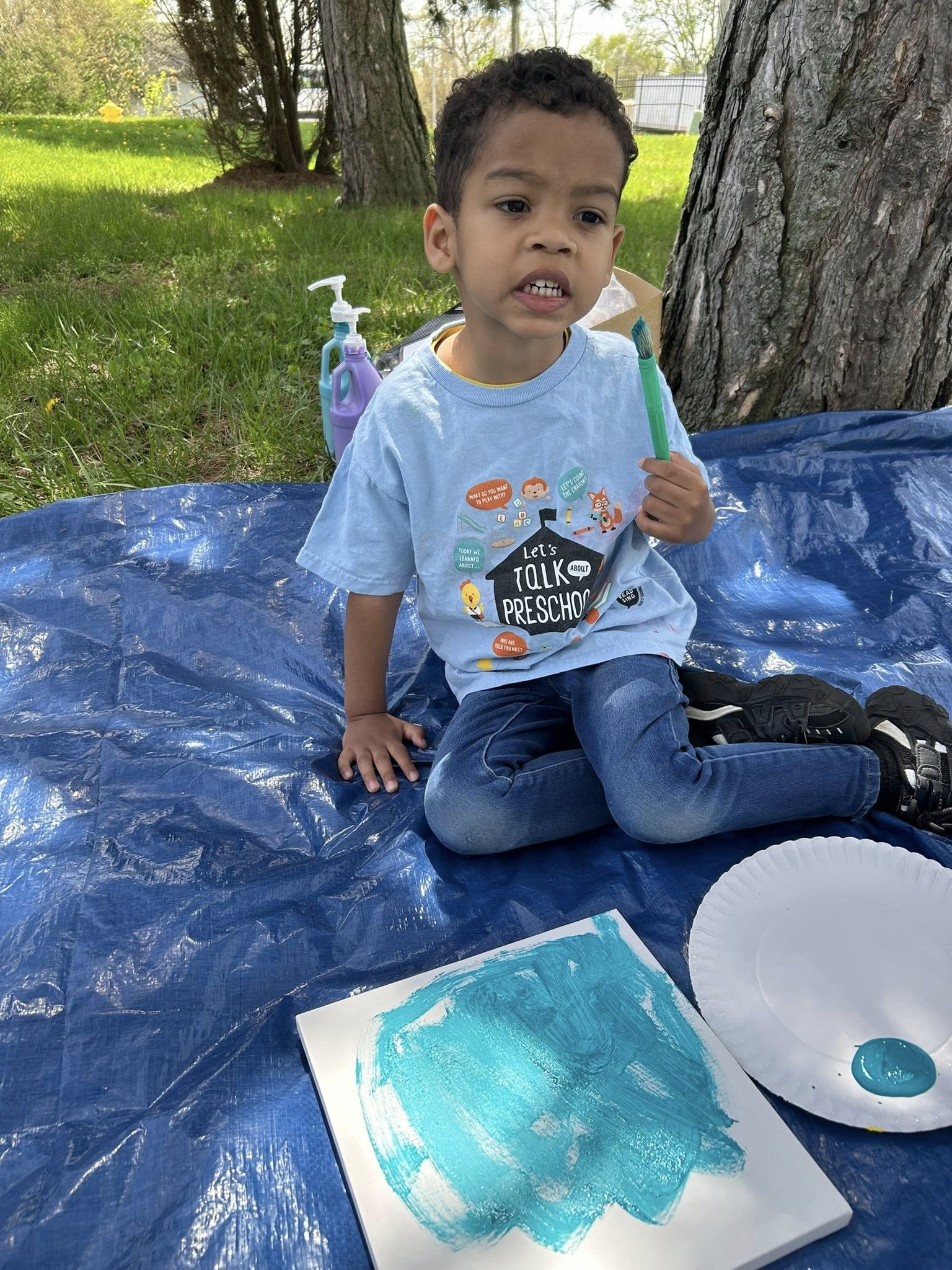 A young boy sitting on a blue tarp outdoors, holding a paintbrush, with a paper plate containing paint, and a canvas with blue painted strokes, surrounded by trees and grass.