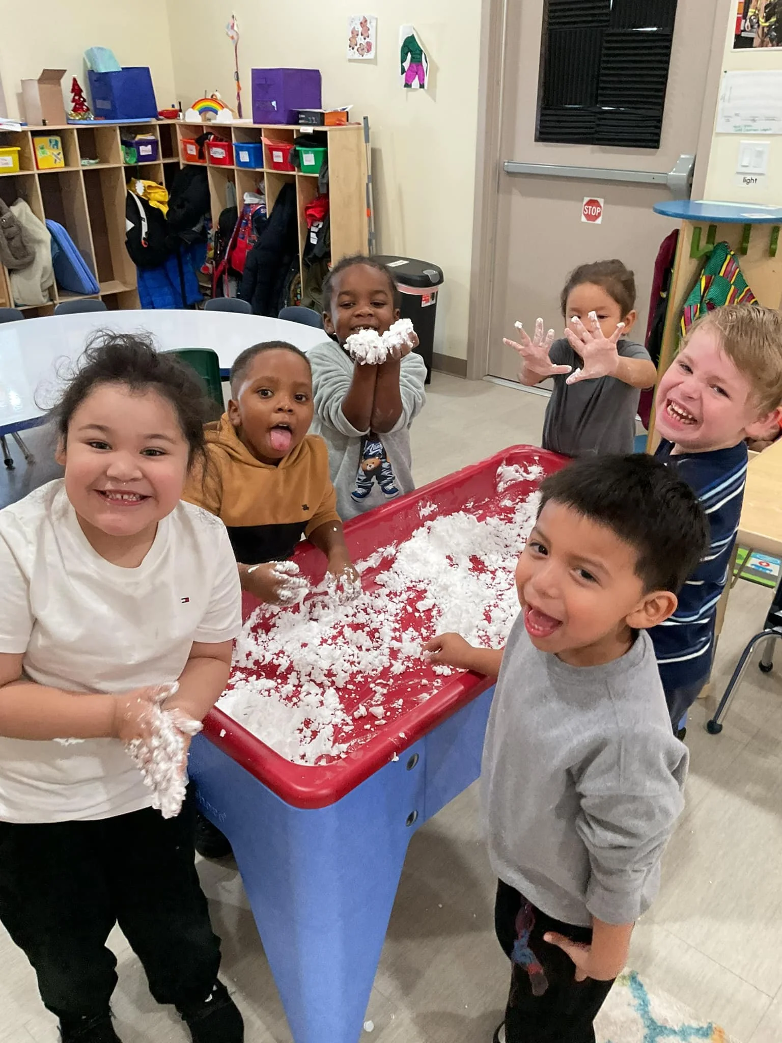 Kids playing with white sensory or craft material at a red and blue table in a classroom.