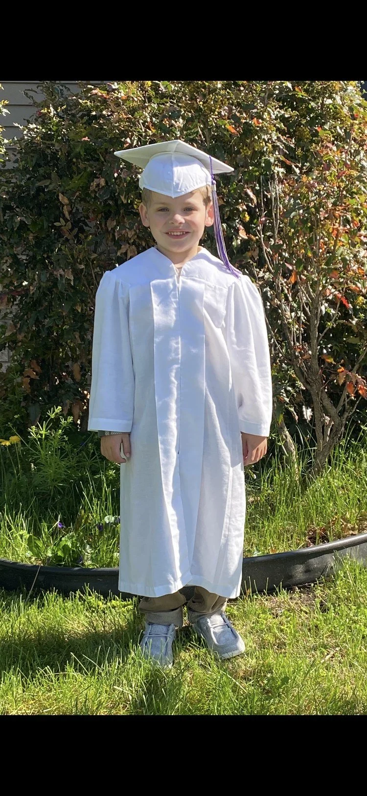 A smiling young boy wearing a white graduation gown and mortarboard cap standing outdoors in front of bushes and grass.