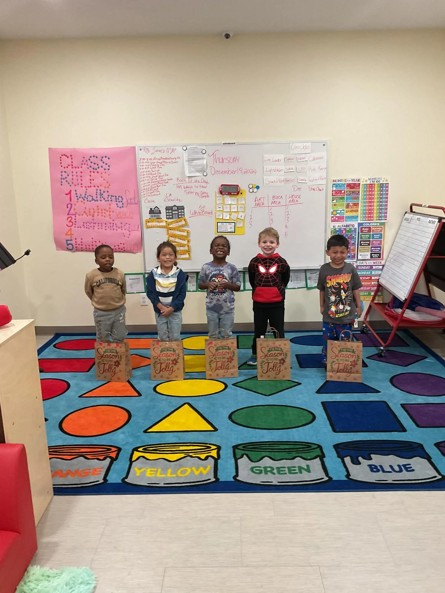 Five young children standing in a classroom in front of a colorful Hopscotch rug, each holding a paper bag with a holiday design. Behind them is a whiteboard with writing and posters, and the classroom appears festive with seasonal decorations.