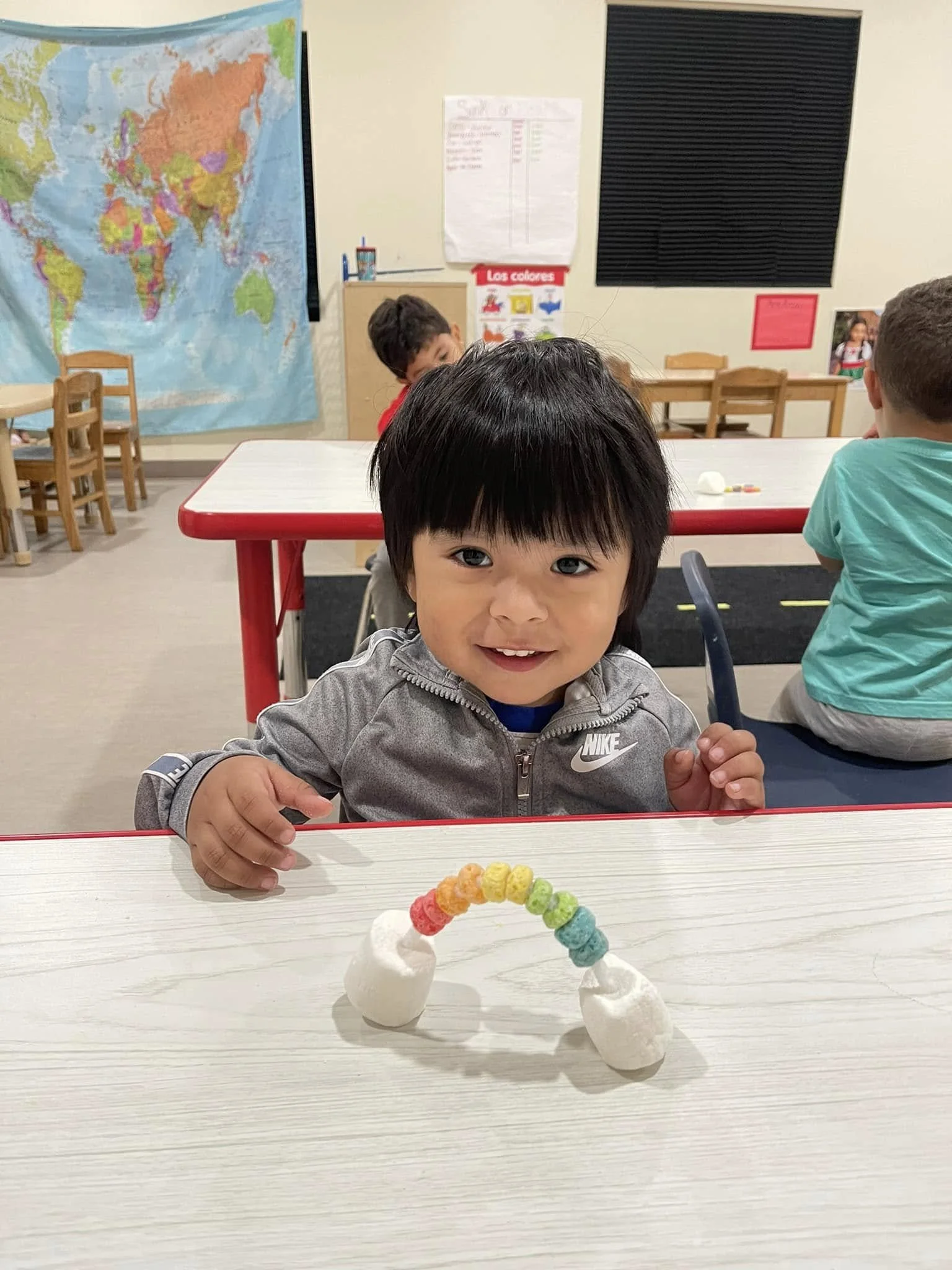 A young boy with black hair and a gray Nike jacket smiling at the camera while sitting at a white table in a classroom. In front of him is a craft made of two marshmallows and colorful cereal pieces arranged in a rainbow shape.