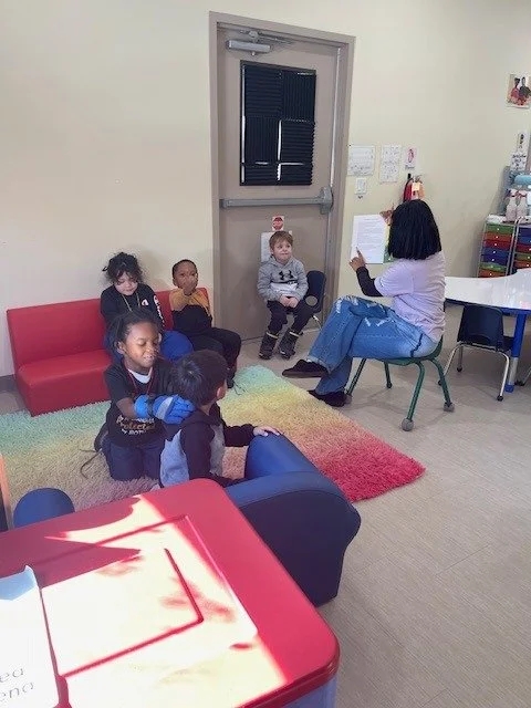 A woman reading a book to five children seated in a room with furniture and a colorful rug.