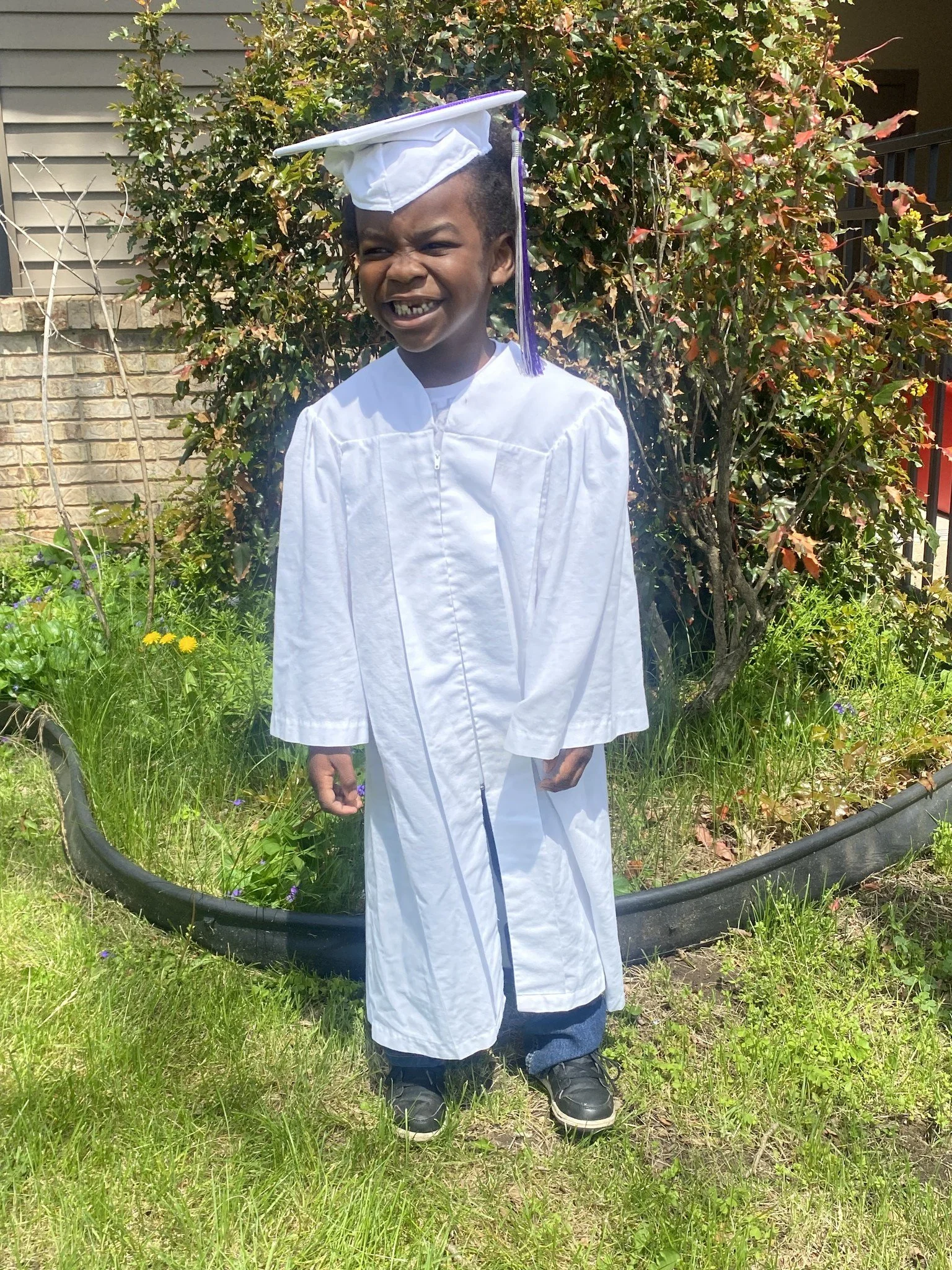 Young boy in a white graduation gown and cap standing outdoors in front of a bush, smiling.