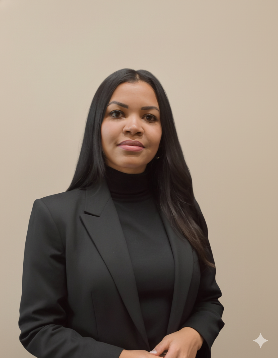 A woman with long dark hair wearing a black blazer and black turtleneck standing against a plain beige wall.