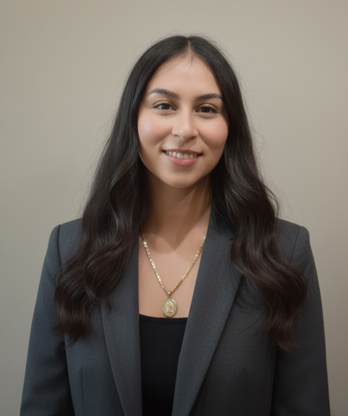 A young woman with long dark wavy hair, smiling, wearing a dark blazer over a black top, and a gold necklace with a pendant, standing against a plain beige wall.