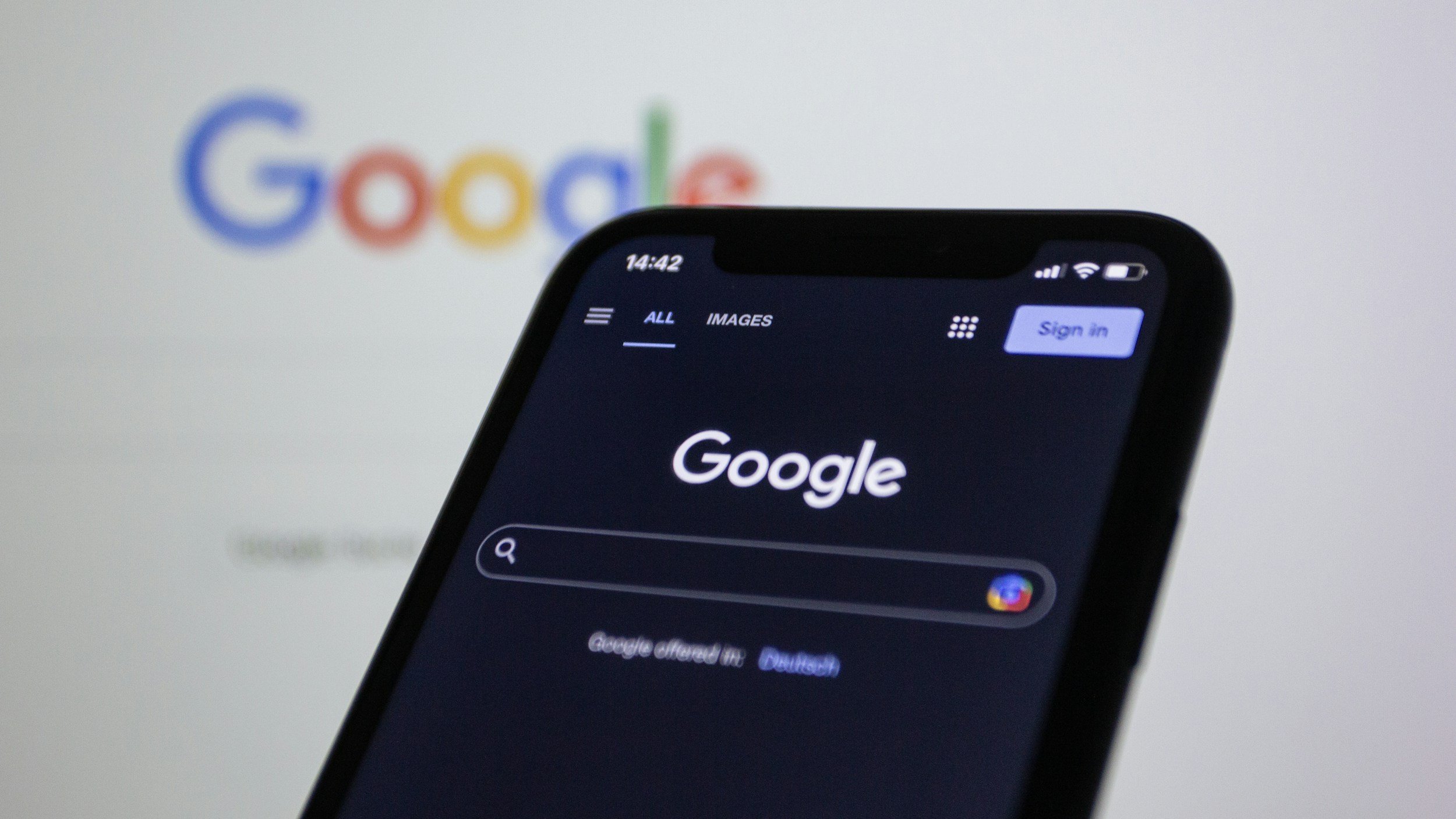 Close-up of a smartphone screen displaying the Google search app with the Google logo visible in the background on a monitor or projected screen.