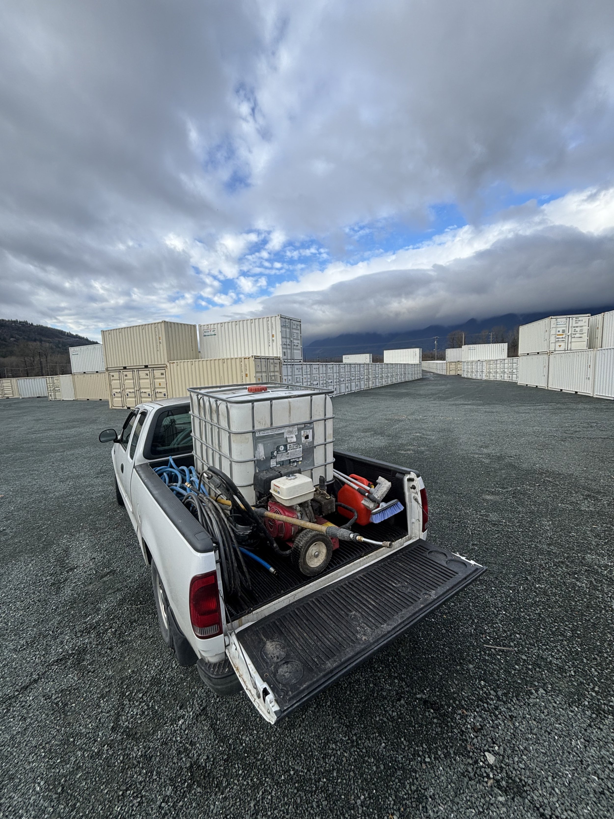 A white pickup truck is parked on a gravel lot filled with shipping containers. The truck's bed contains a large water tank, pressure washer, hoses, and cleaning tools. The sky is cloudy with patches of blue.