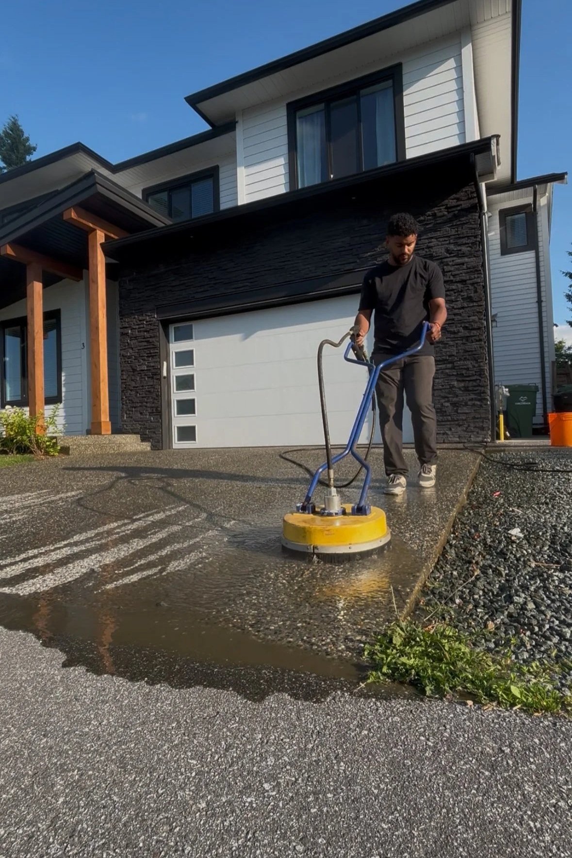 A man is cleaning a concrete driveway with a power surface cleaner, water is flowing from the machine, and he is wearing a black shirt, gray pants, and sneakers outside a modern house.
