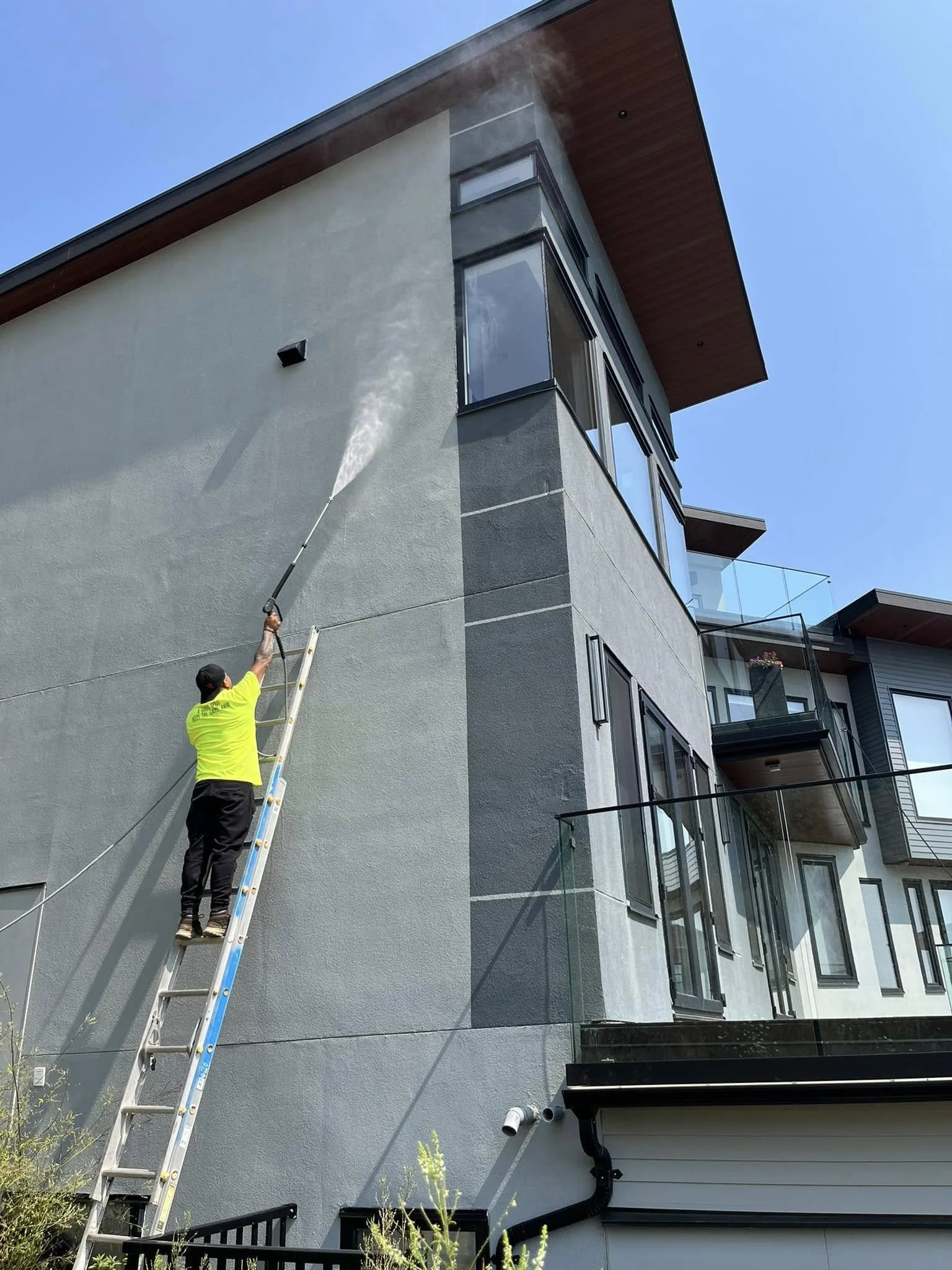 A person standing on a ladder cleaning or spraying the exterior wall of a modern multi-story building with a pressure washer or sprayer.