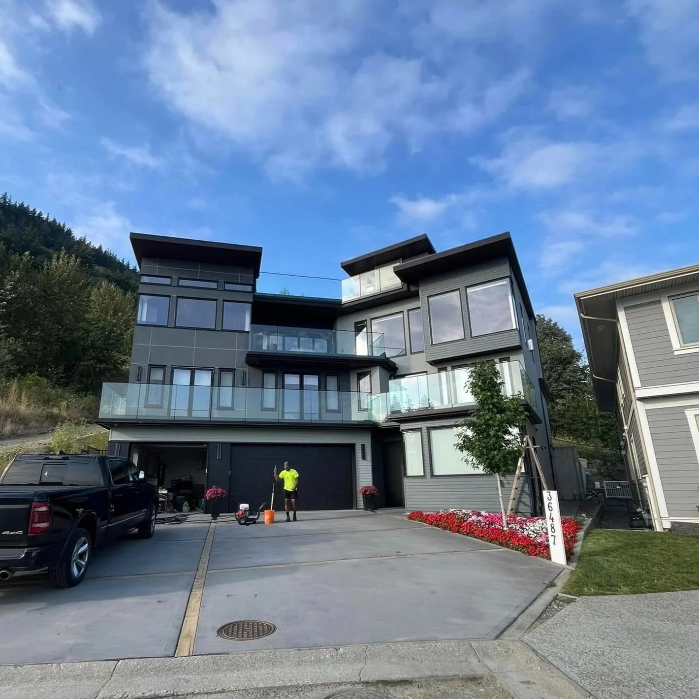 Modern multi-story gray house with large windows and glass balcony railings, parked black pickup truck, person in yellow shirt standing with cleaning equipment, landscaped flower bed with red and pink flowers, and neighboring house on the right, under a blue sky with clouds.