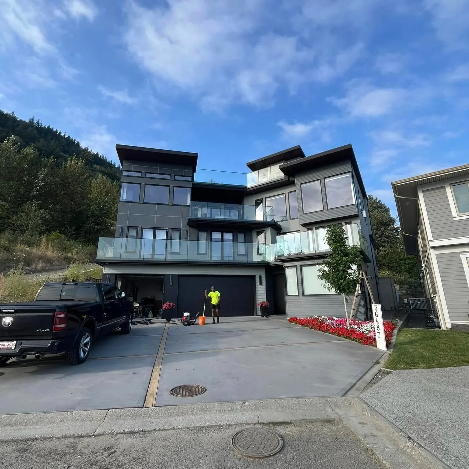 Modern multi-story house with gray exterior, glass balconies, and large windows. A man in a neon yellow shirt stands in the driveway near parked cars, with a blue sky with some clouds above.
