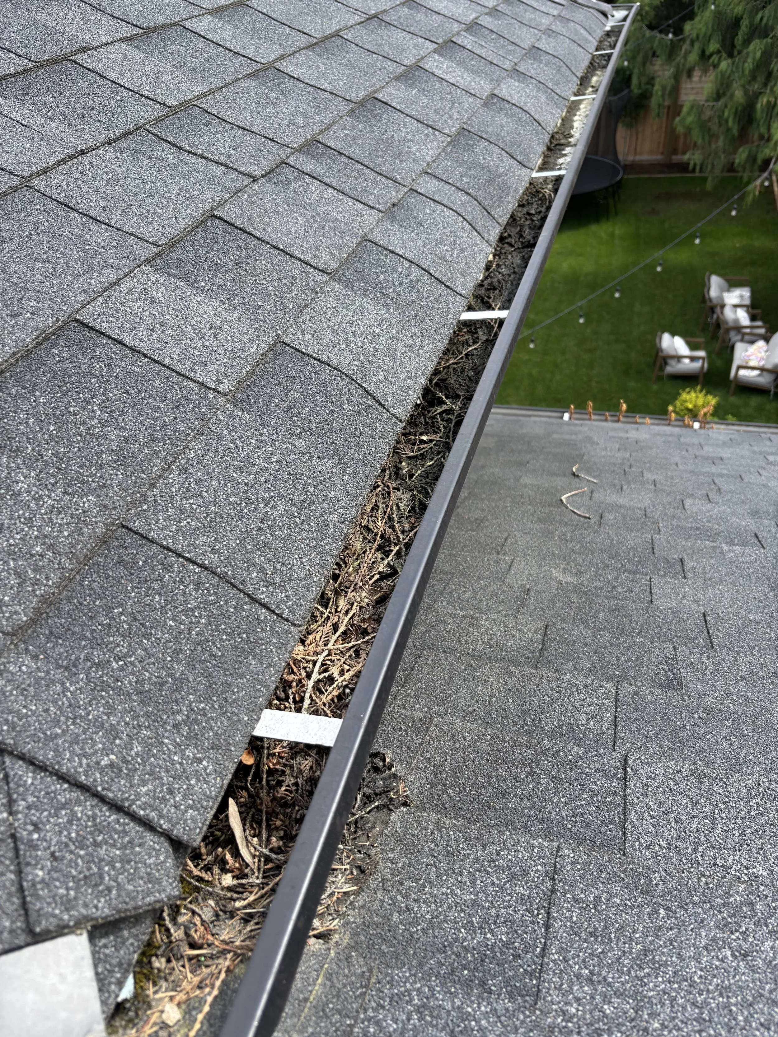 Close-up of a roof with gray asphalt shingles and a gutter filled with dirt, debris, and leaves.