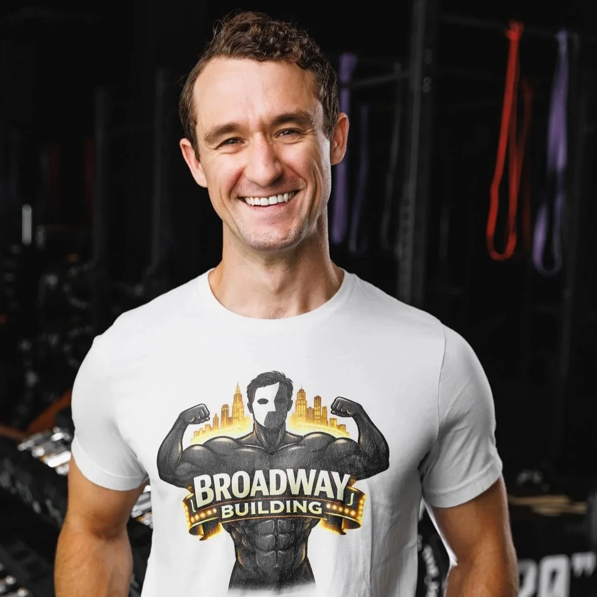 A smiling man with short brown hair wearing a white T-shirt with a graphic of a muscular man and the words 'Broadway Building' on it. He is standing in a gym or workout area with various fitness equipment in the background.