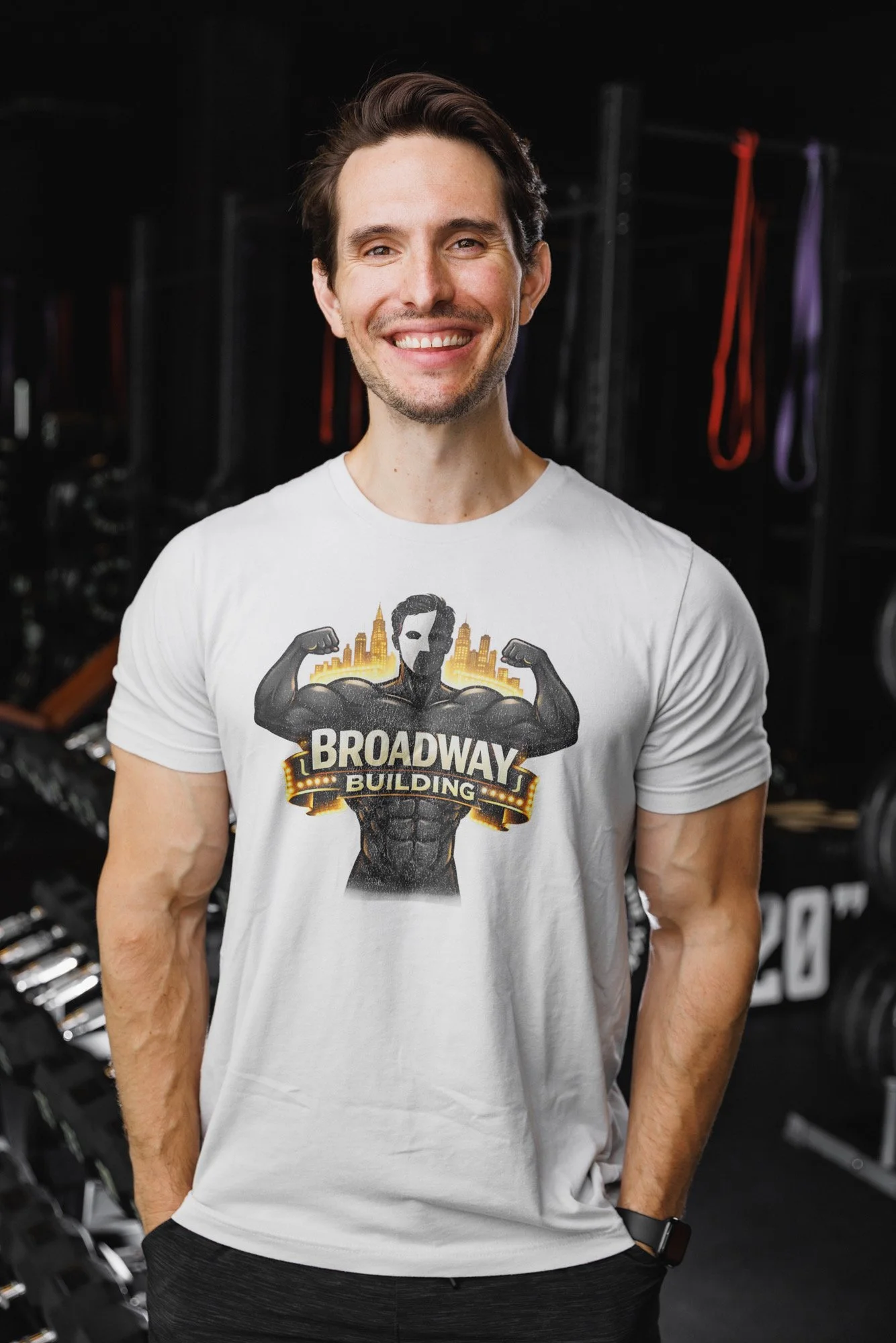 A smiling man wearing a white T-shirt with a graphic of a muscular superhero figure and the words BROADWAY BUILDING, standing in a gym with workout equipment and resistance bands in the background.