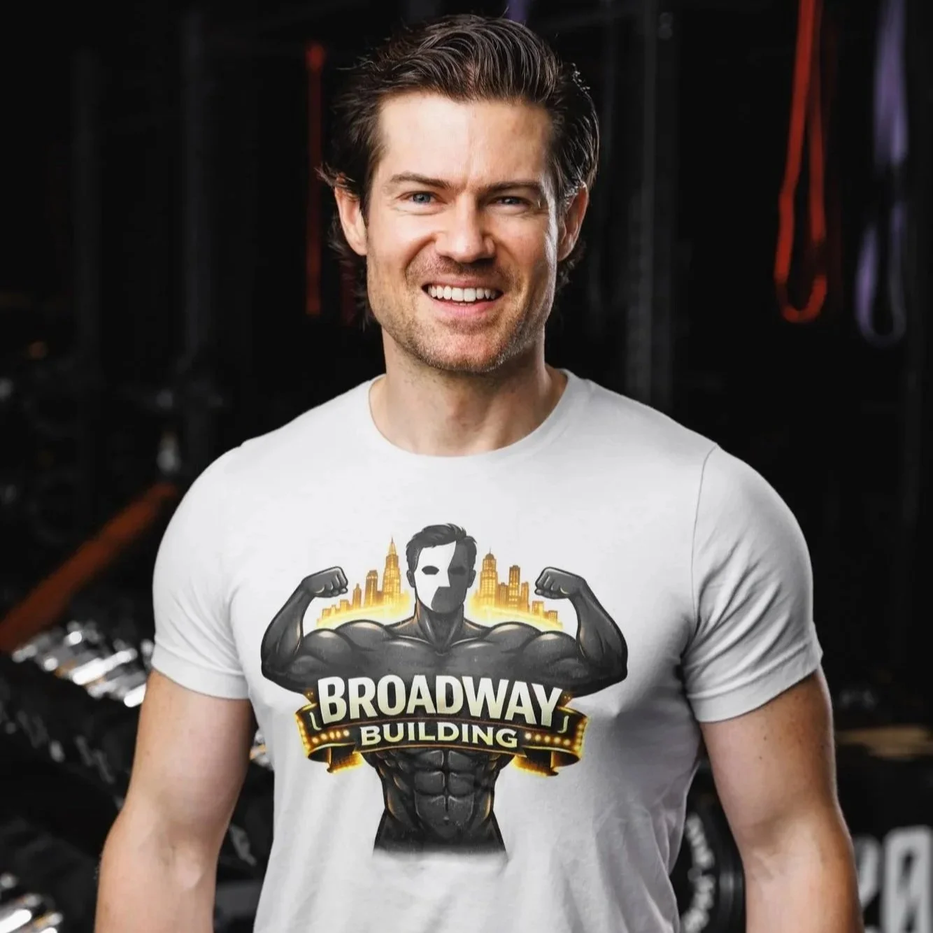 A smiling man with dark brown hair and a light beard, wearing a white T-shirt with a muscular cartoon figure and the text "Broadway Building" on it, standing in a gym with workout equipment in the background.