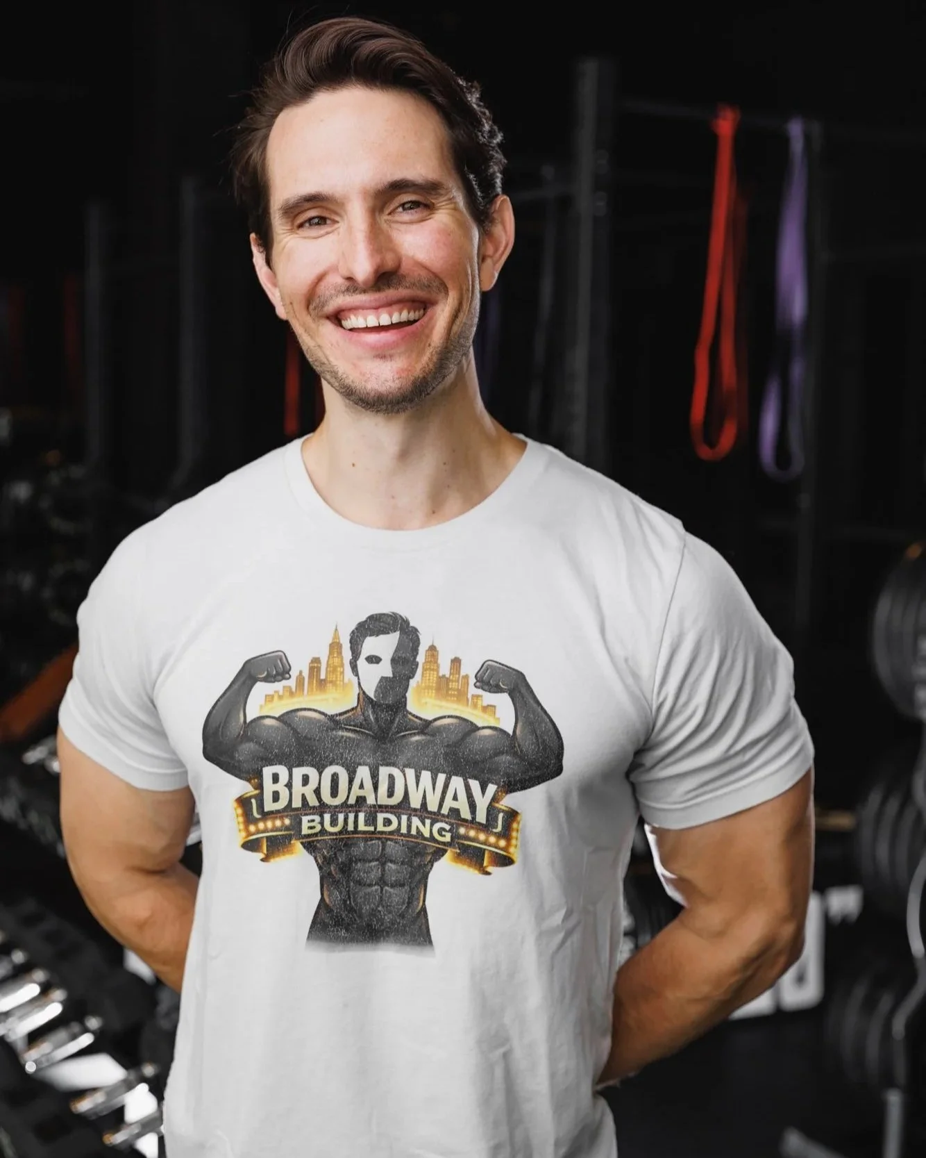 Man smiling, wearing a white T-shirt with a graphic design of a muscular superhero figure with flexed arms and the words 'Broadway Building' on it, standing in front of gym equipment and resistance bands.