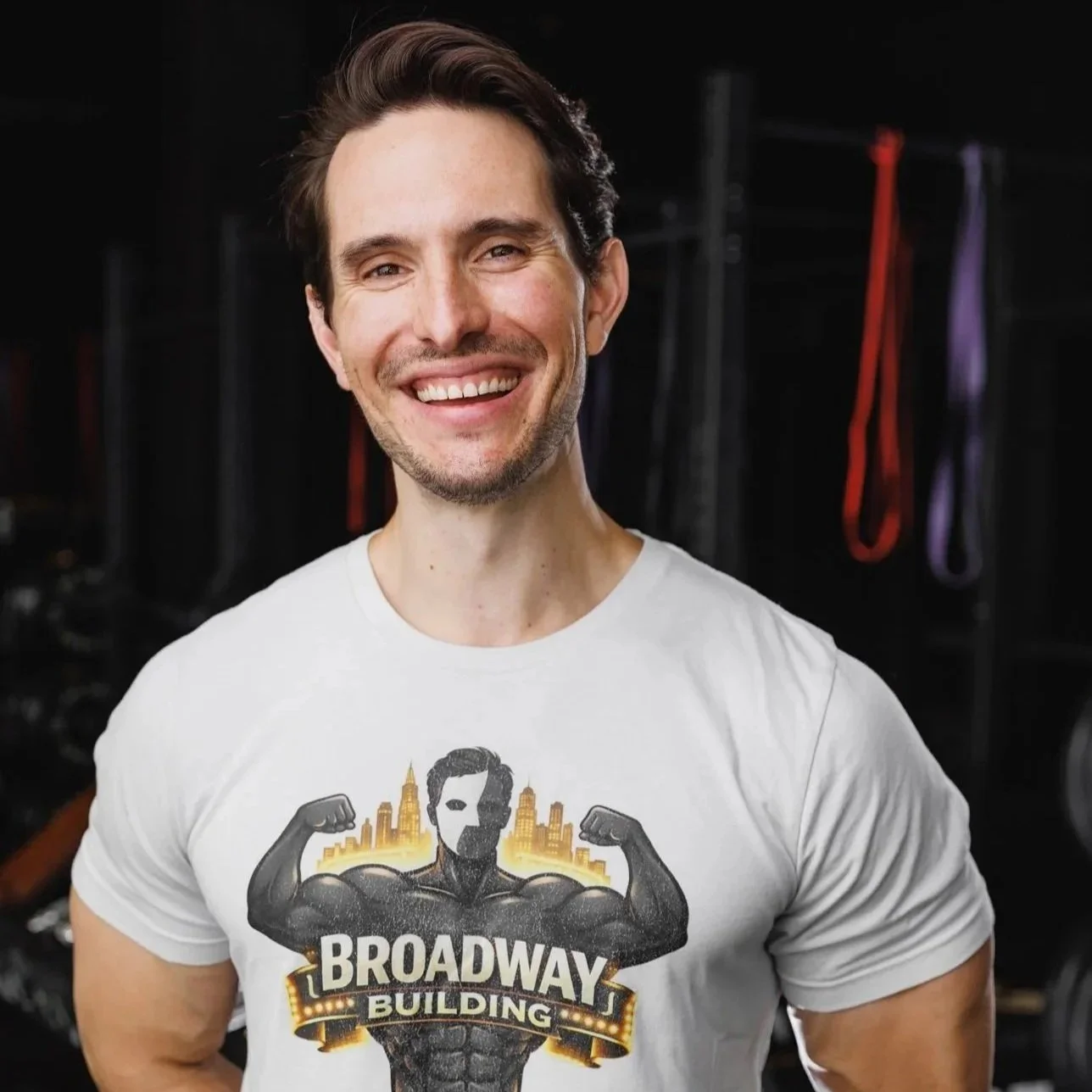 A happy man with dark hair and a beard smiling in front of a black background with exercise rings hanging, wearing a white T-shirt featuring a muscular figure with the text 'Broadway Building' and a city skyline.